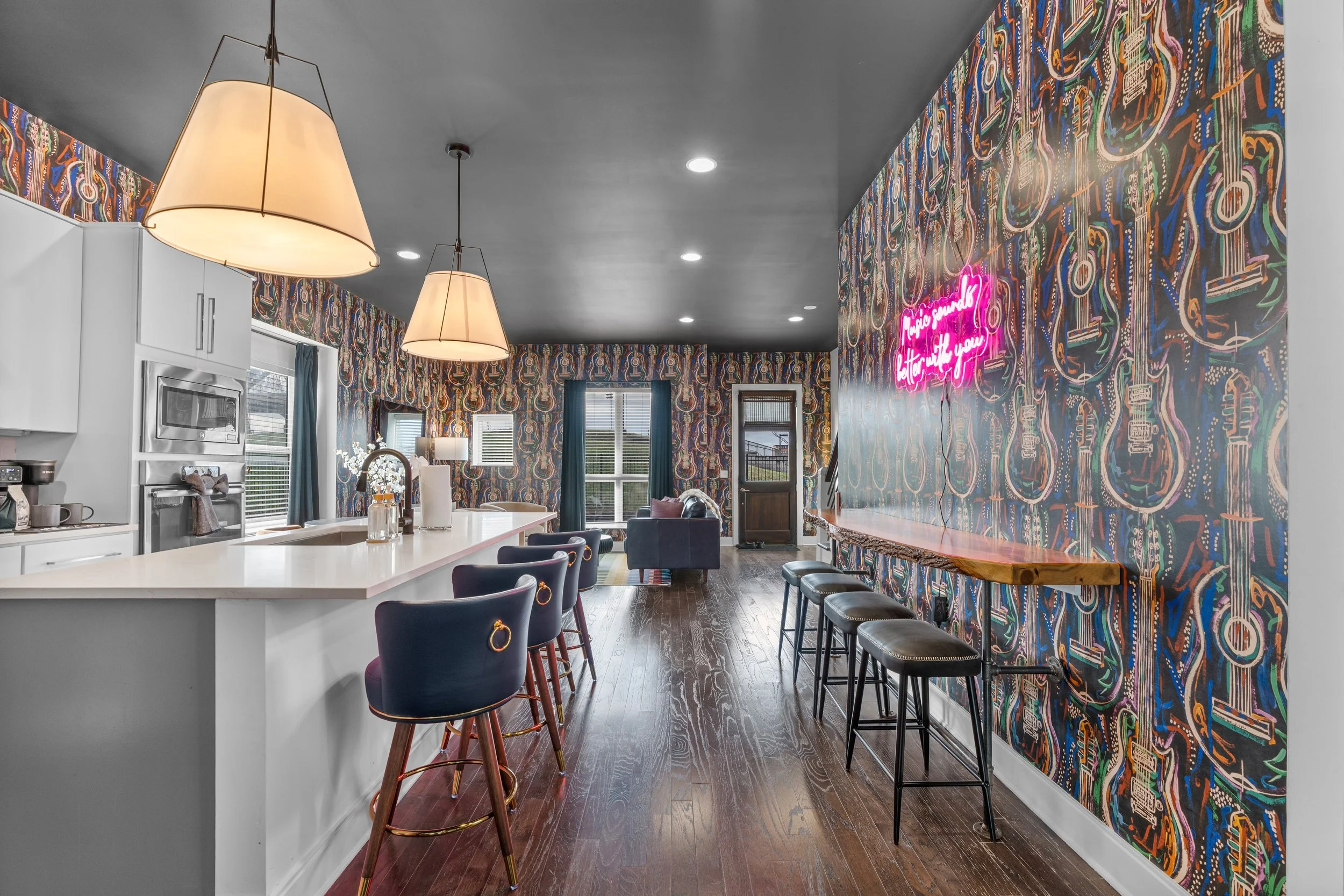 Interior of a modern kitchen and living area with a colorful patterned accent wall, white cabinetry, a white island with barstools, pendant lights, and a neon sign on the wall that says 'Music sounds better with you'.