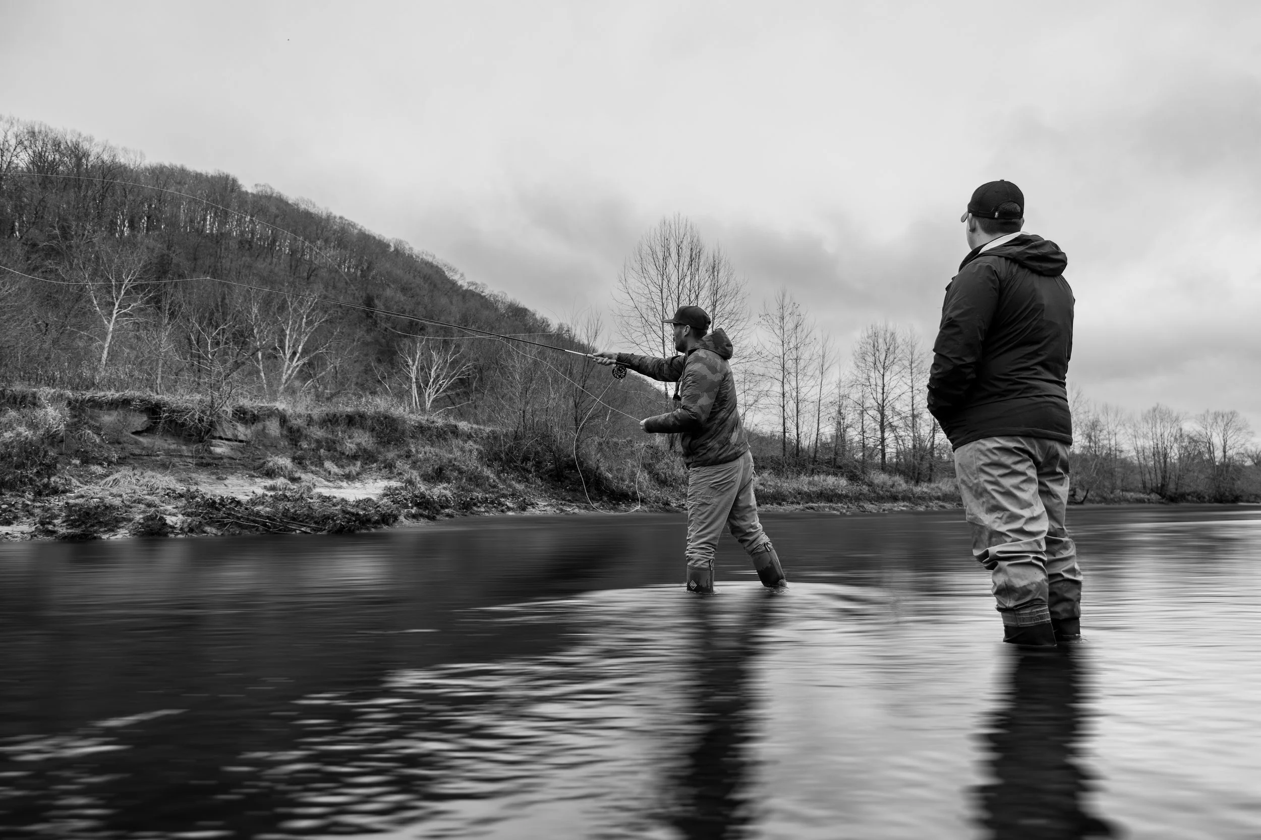 Two men fishing in a river, one casting a line while the other watches, with trees and a hilly landscape in the background, in a black and white photograph.