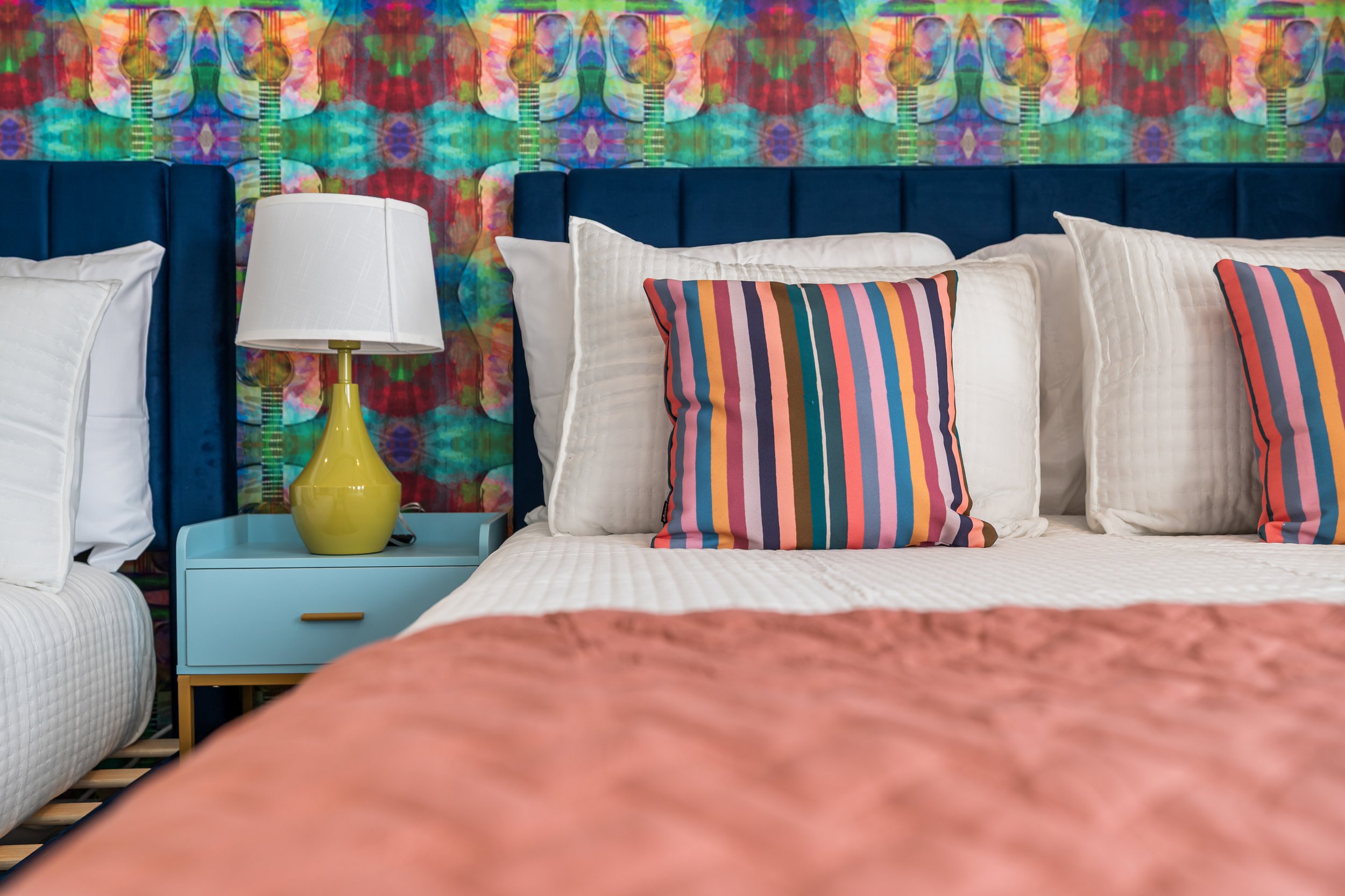 A bedroom with a colorful, tie-dye patterned wallpaper, a blue upholstered headboard, and a white bed with white and multicolored striped pillows. There is a yellow bedside lamp on a blue nightstand.