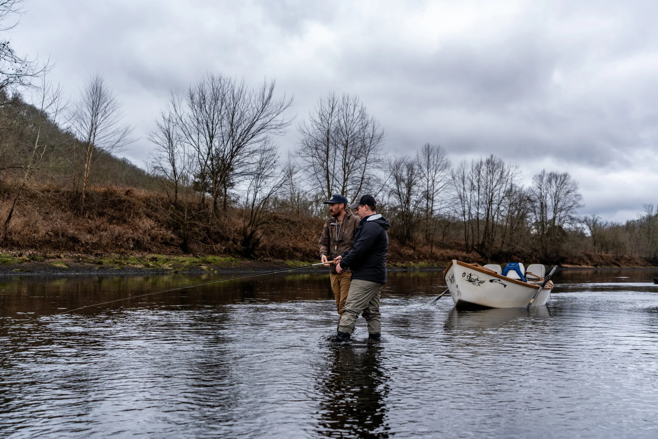 Two men are fishing in a river with a small boat nearby. The sky is overcast, and the background features leafless trees and a hillside.