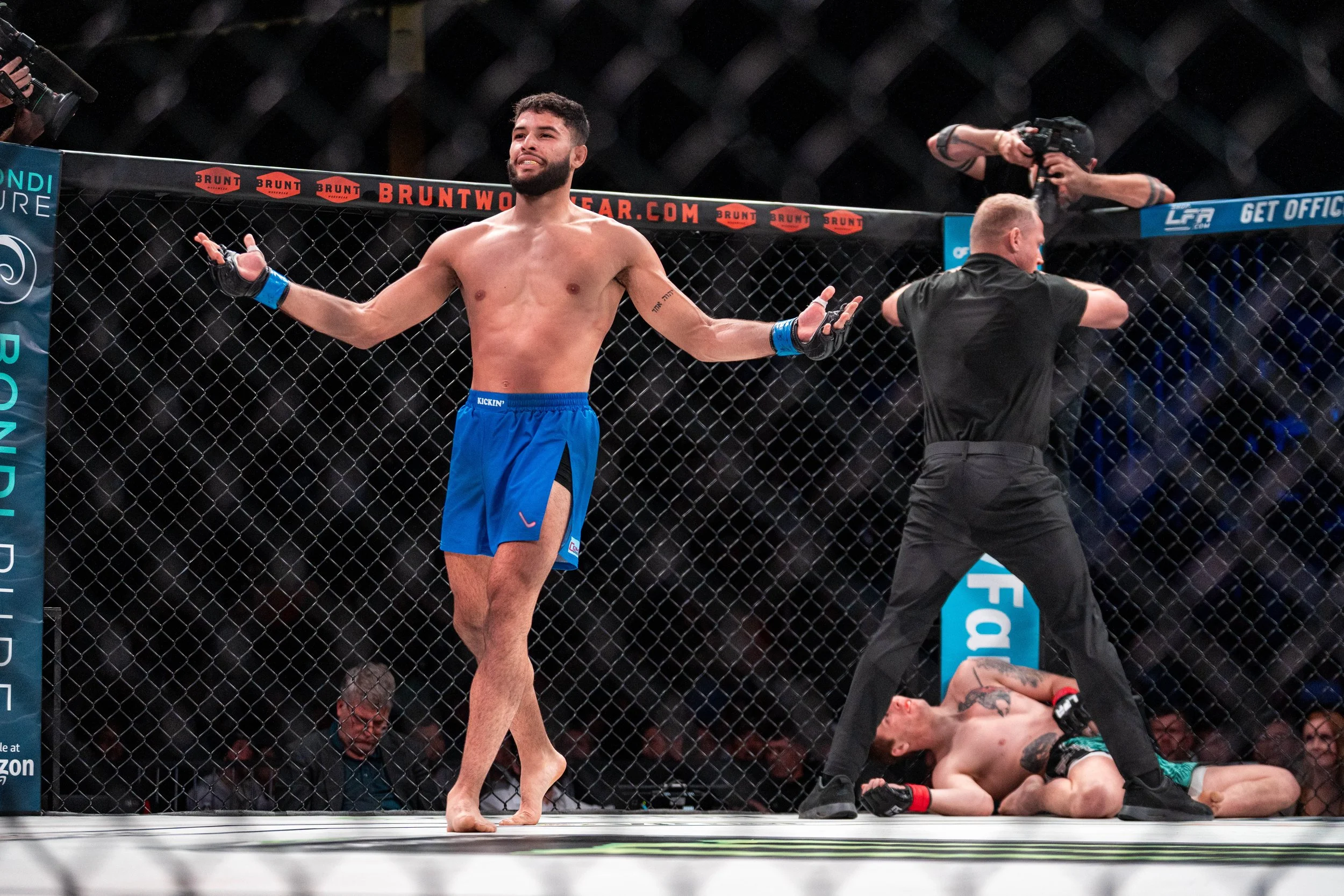 Mixed martial arts fight inside a cage, with one fighter standing with arms spread and another fighter lying on the ground, possibly knocked out, while a referee watches closely.