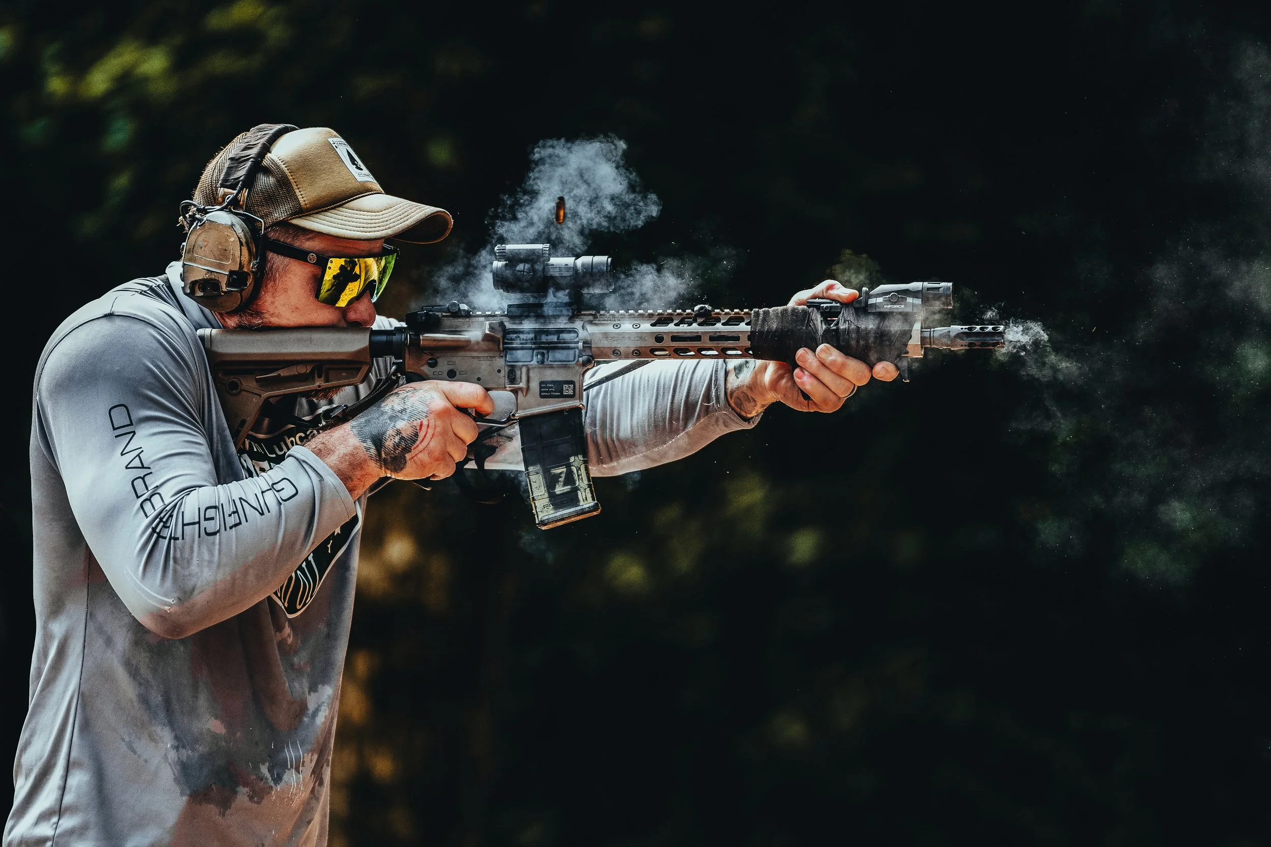 A man wearing protective gear, including a cap, sunglasses, and headphones, is aiming and firing a tactical rifle outdoors as smoke and a bullet are visible.