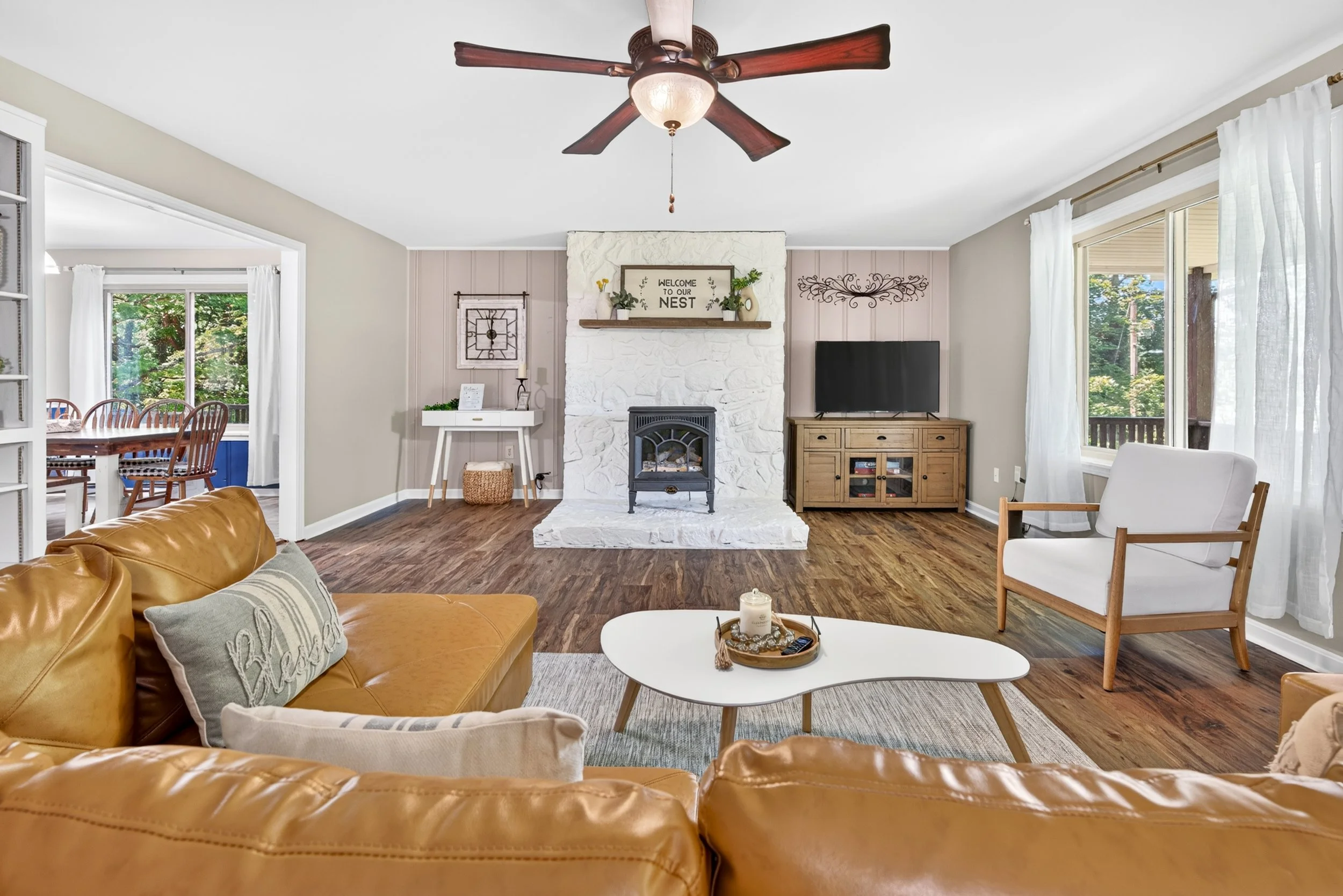 Living room with a tan leather sofa, gray and white pillows, white coffee table, small round tray with candles, a wooden armchair with white cushion, large windows with white curtains, wood floor, fireplace with mantel decorated with plants and a sig
