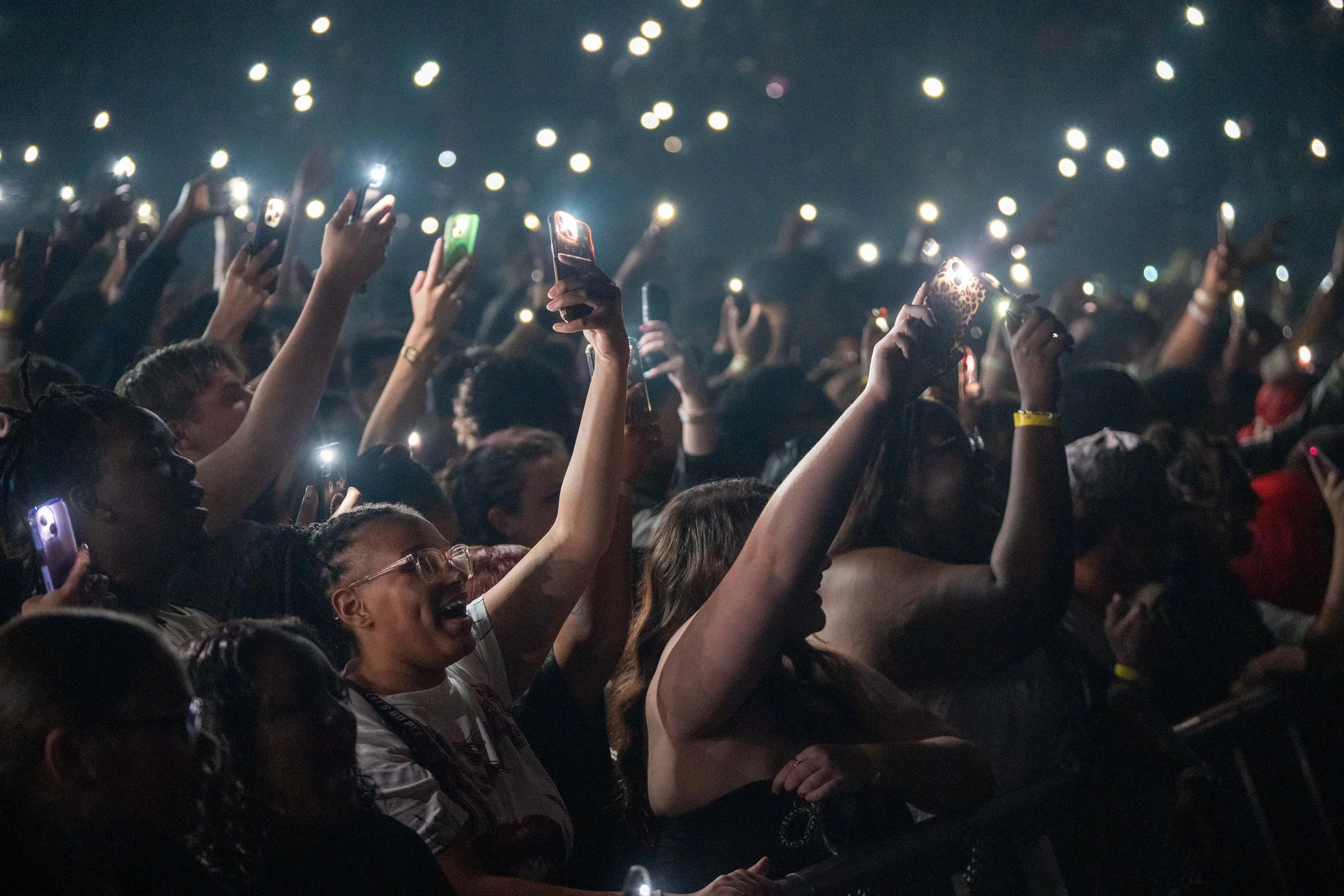 A crowd of people at a concert or event, holding up their phones with flashlight features turned on, creating a sea of lights in the dark.