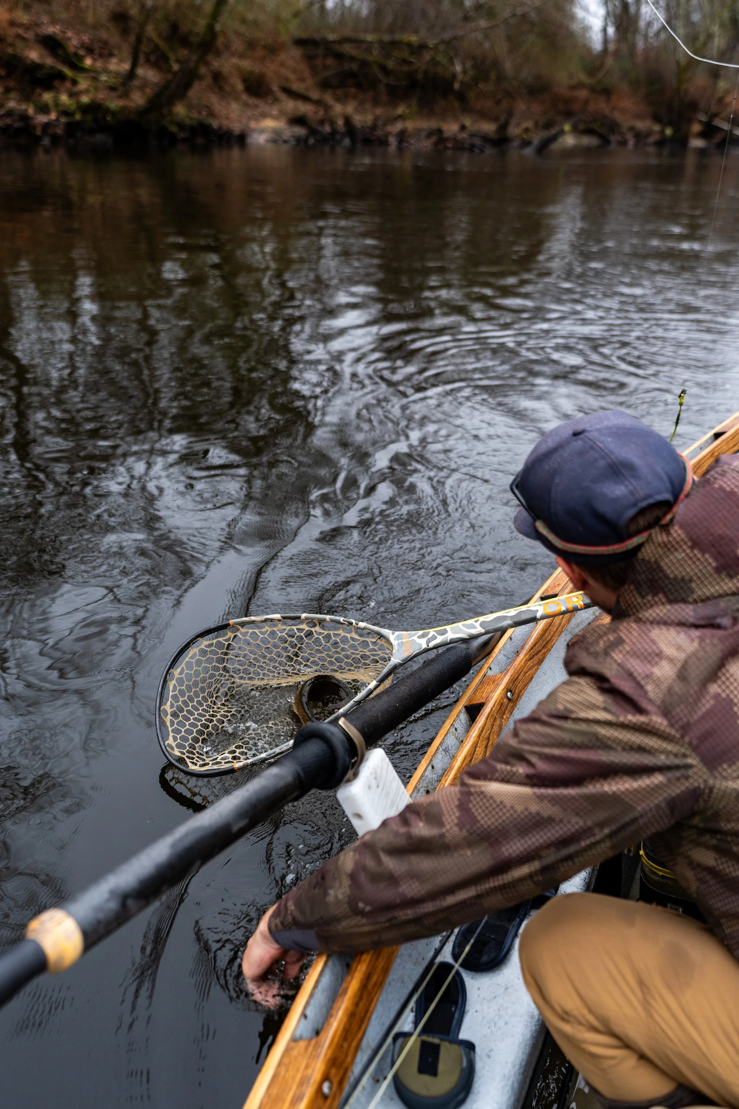 Person wearing a cap and brown jacket catching a fish with a fishing net while sitting in a boat on a river surrounded by trees.