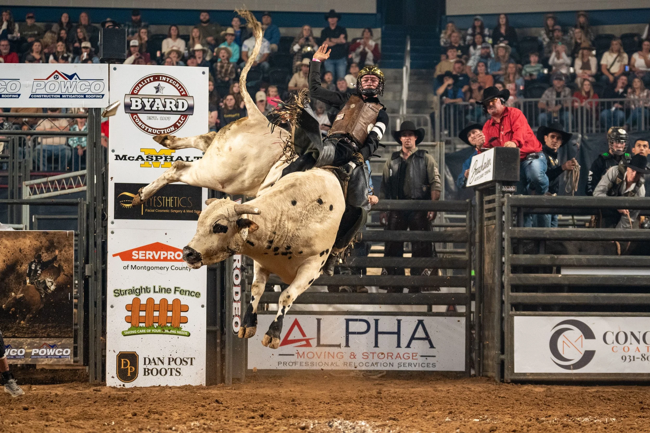 A rodeo rider wearing a helmet and protective vest riding a bucking bull during a rodeo event, with an audience watching in the background.