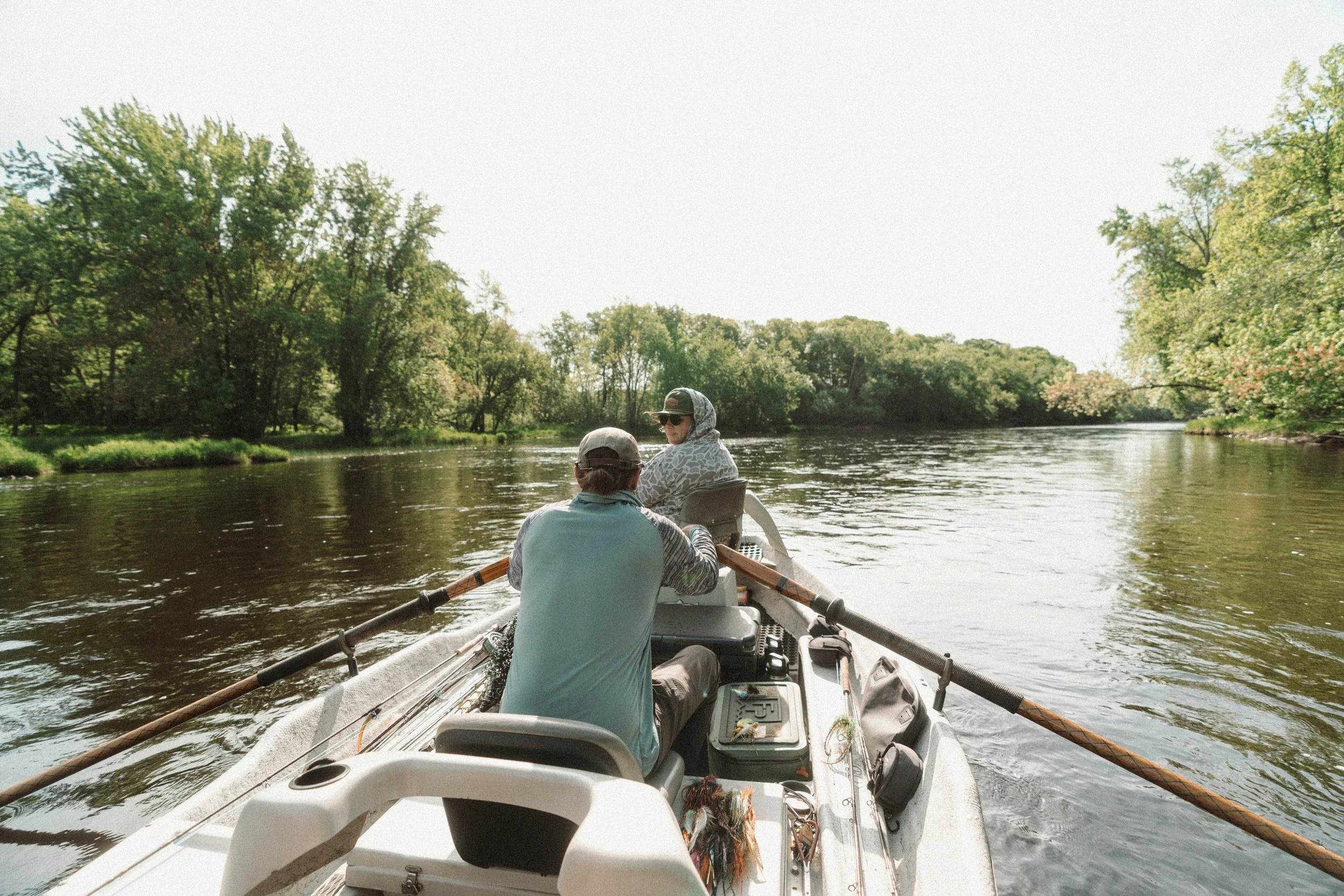 Two people are sitting in a small boat on a river surrounded by green trees. One person is facing away and the other is wearing sunglasses and a hoodie. The boat has oars and some gear inside.