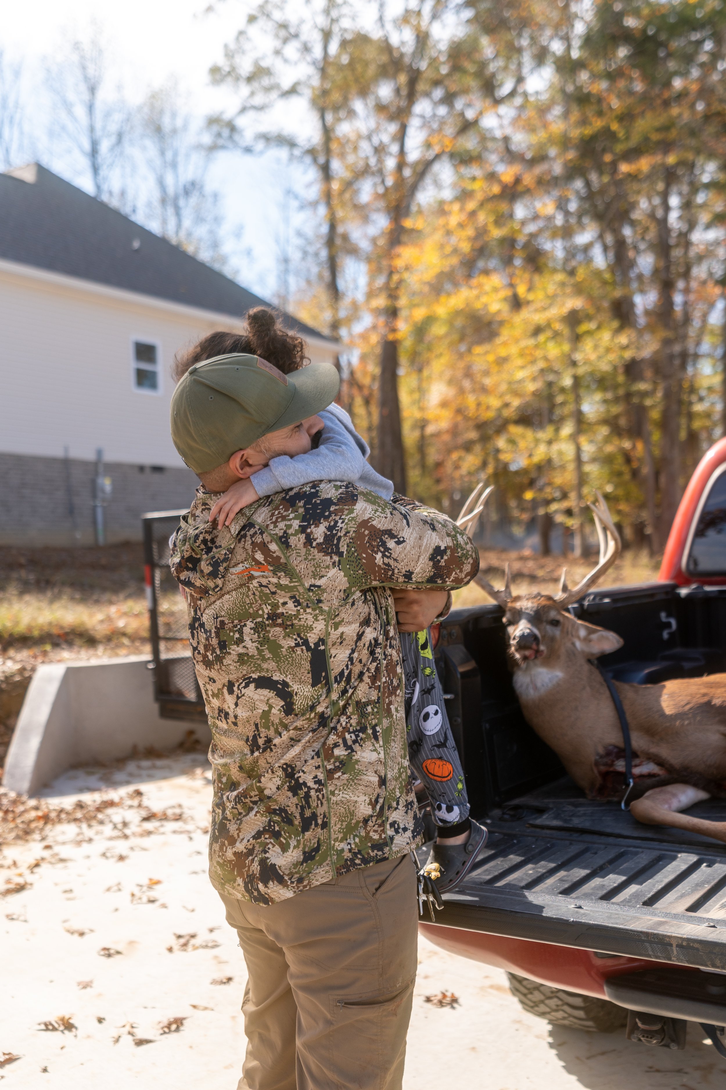 A man wearing a camouflage jacket and tan pants is hugging a young girl in a gray hoodie outdoors during autumn. The girl has curly hair and is wearing a green cap. A deer is sitting in the back of a pickup truck nearby. The background features a hou