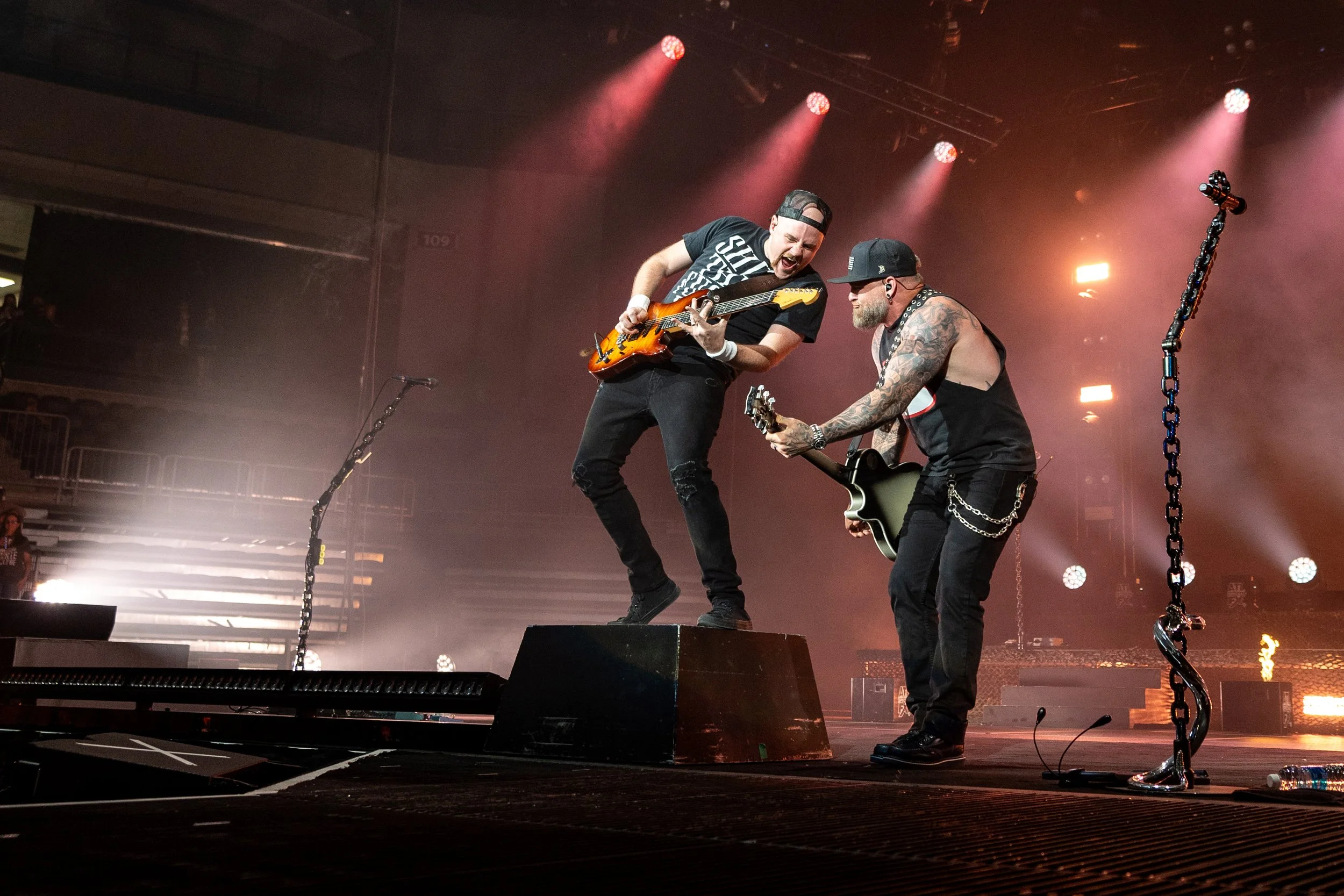 Two musicians on stage, one playing an electric guitar and the other a microphone, with colorful stage lights and smoke effects in the background.