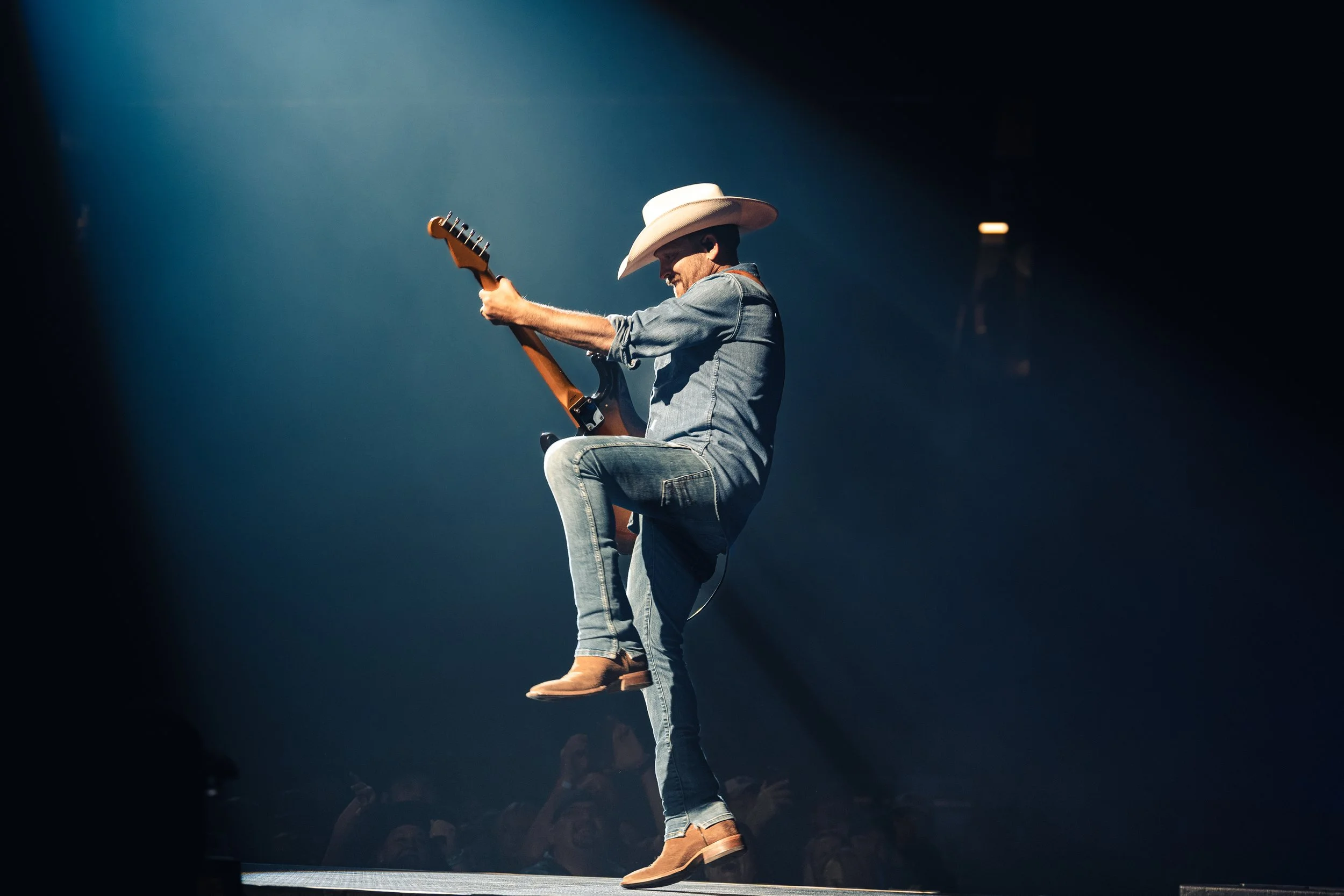 A man wearing a cowboy hat, jeans, and boots is playing an electric guitar on stage with a spotlight illuminating him. He is standing with one foot lifted and a dark background surrounding him.