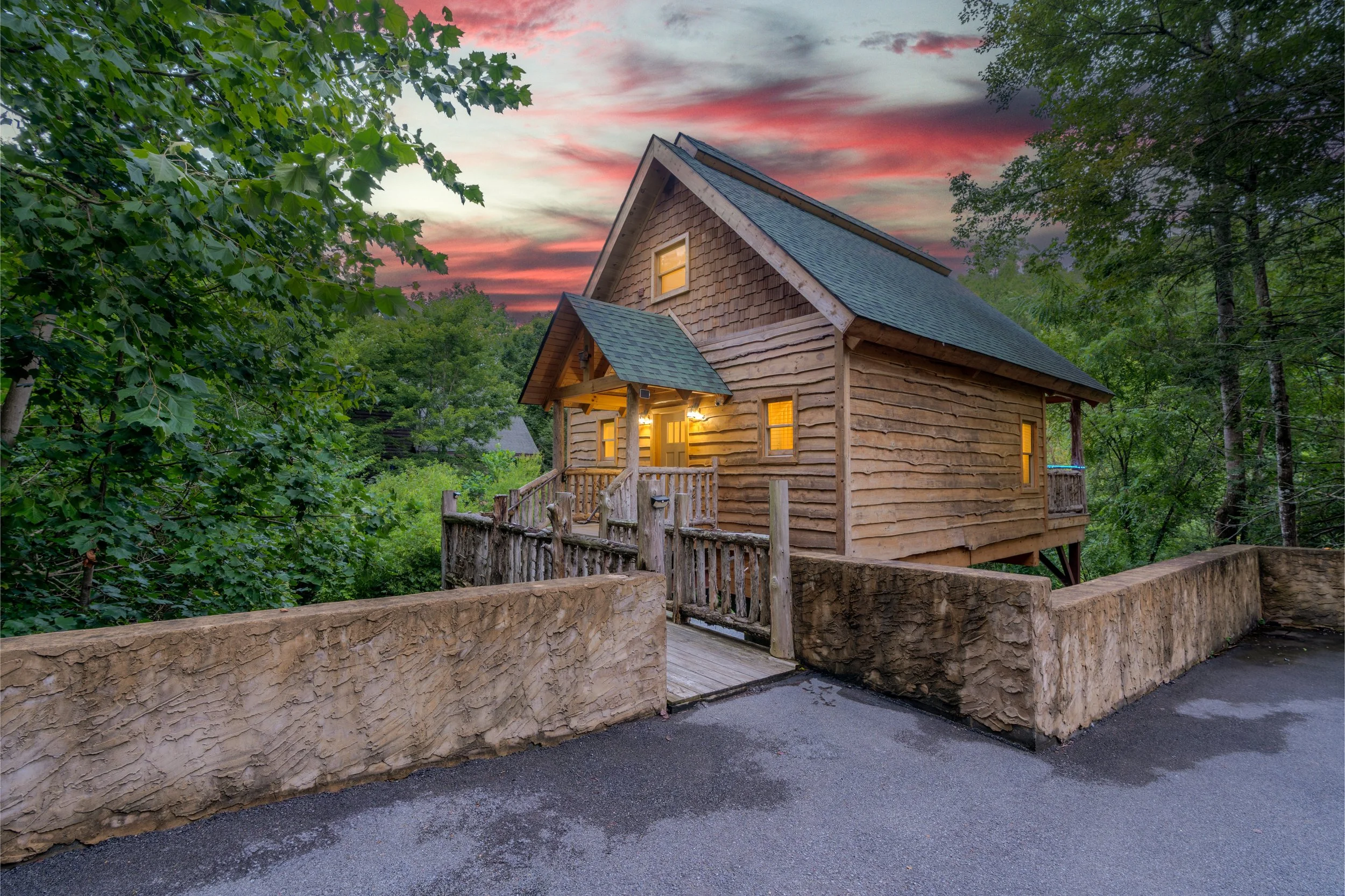 A small wooden house with a steep roof and lit windows, surrounded by green trees at sunset, with a stucco fence and paved driveway in the foreground.
