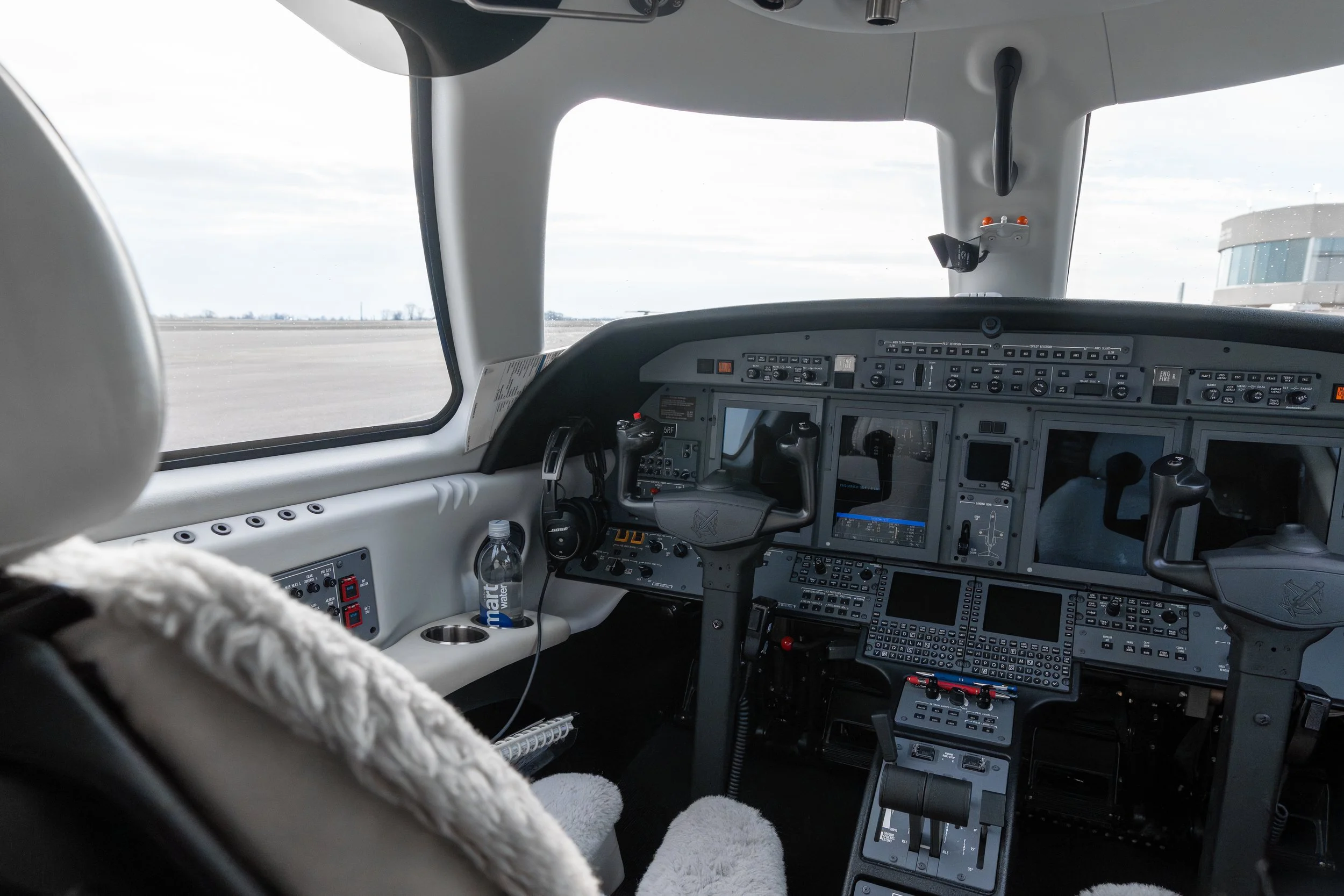 Inside view of a small airplane cockpit with multiple flight instruments, controls, and screens, with a view of an airport runway through the front window.