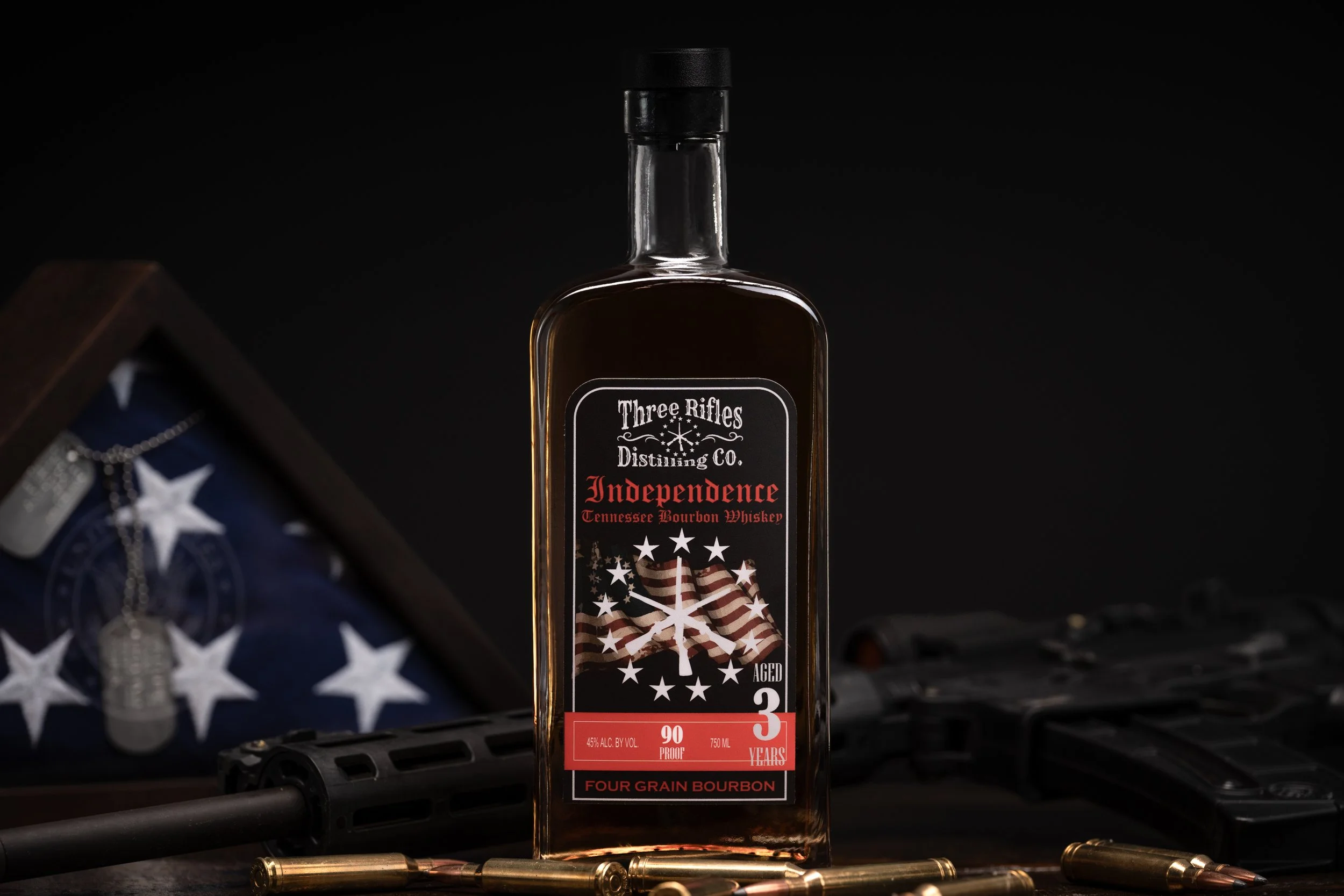 A bottle of Three Ridges Distilling Co. Independence Tennessee Bourbon whiskey with a label featuring an American flag design, set against a dark background with ammunition rounds in the foreground.