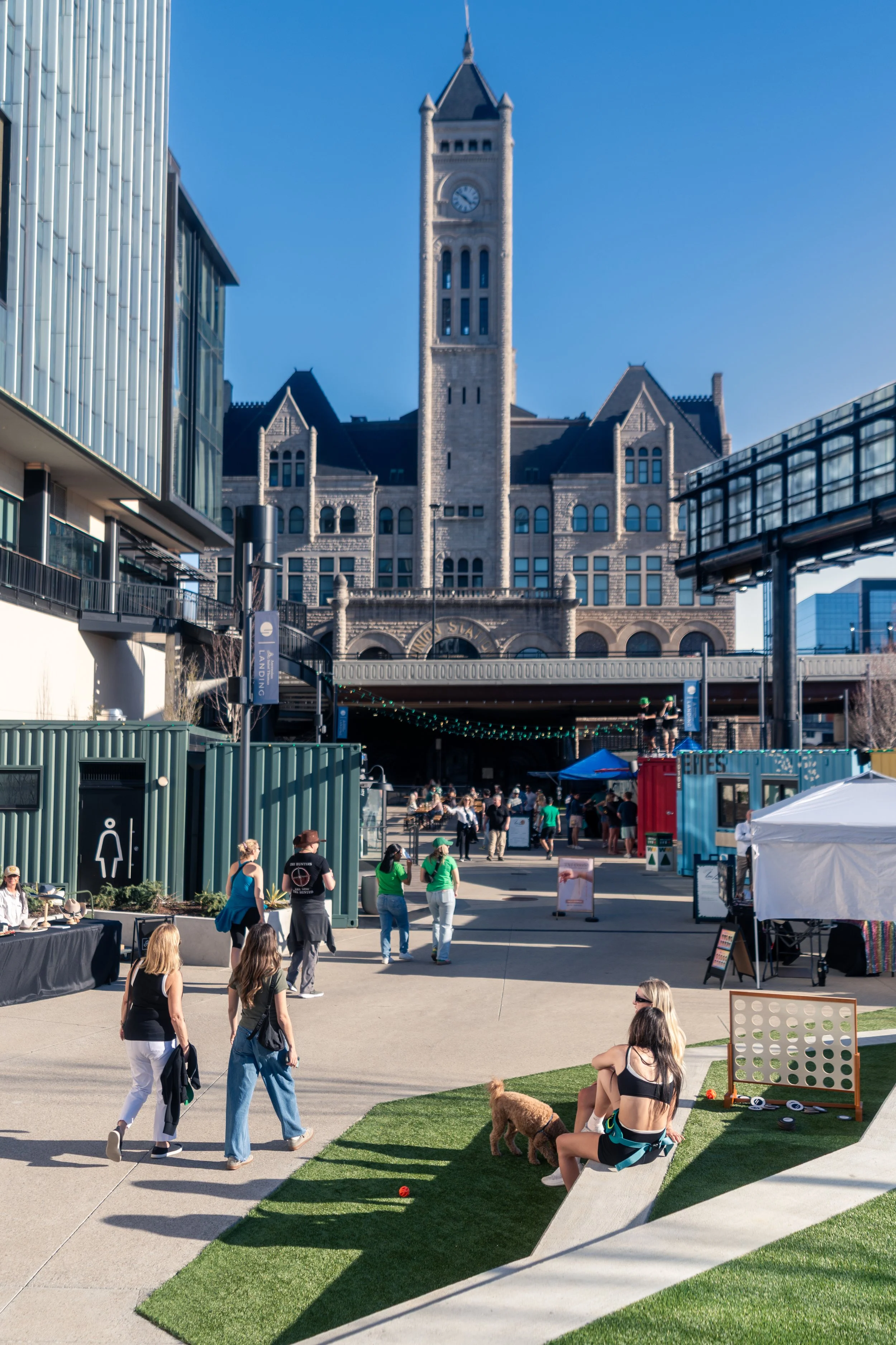 People gathered at an outdoor event in front of a historic building with a clock tower, some sitting on the grass and others walking or talking, with vendor tents and a small dog present.