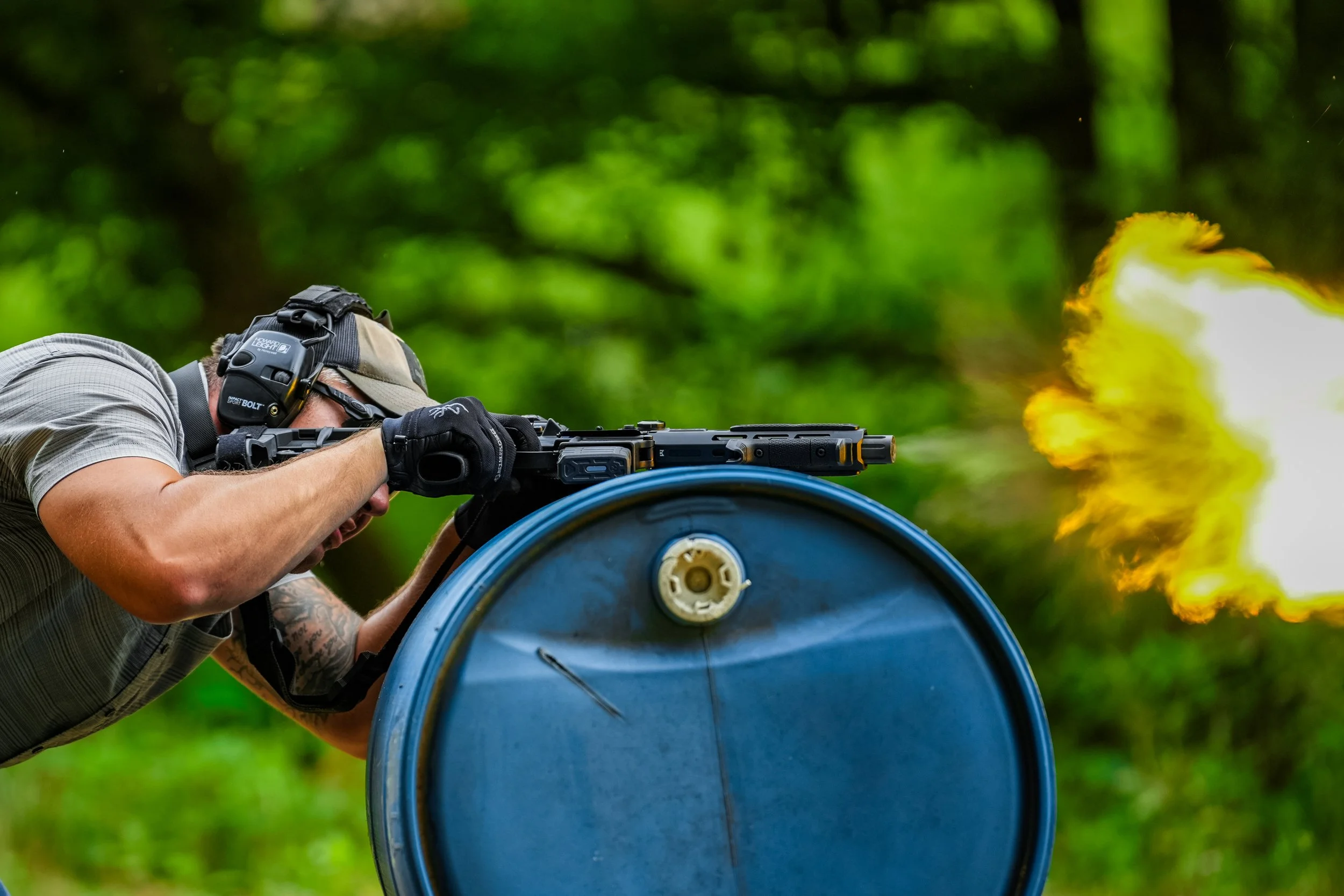 A man in a cap and gloves aiming a firearm out of a tube, with a muzzle flash coming from the gun.