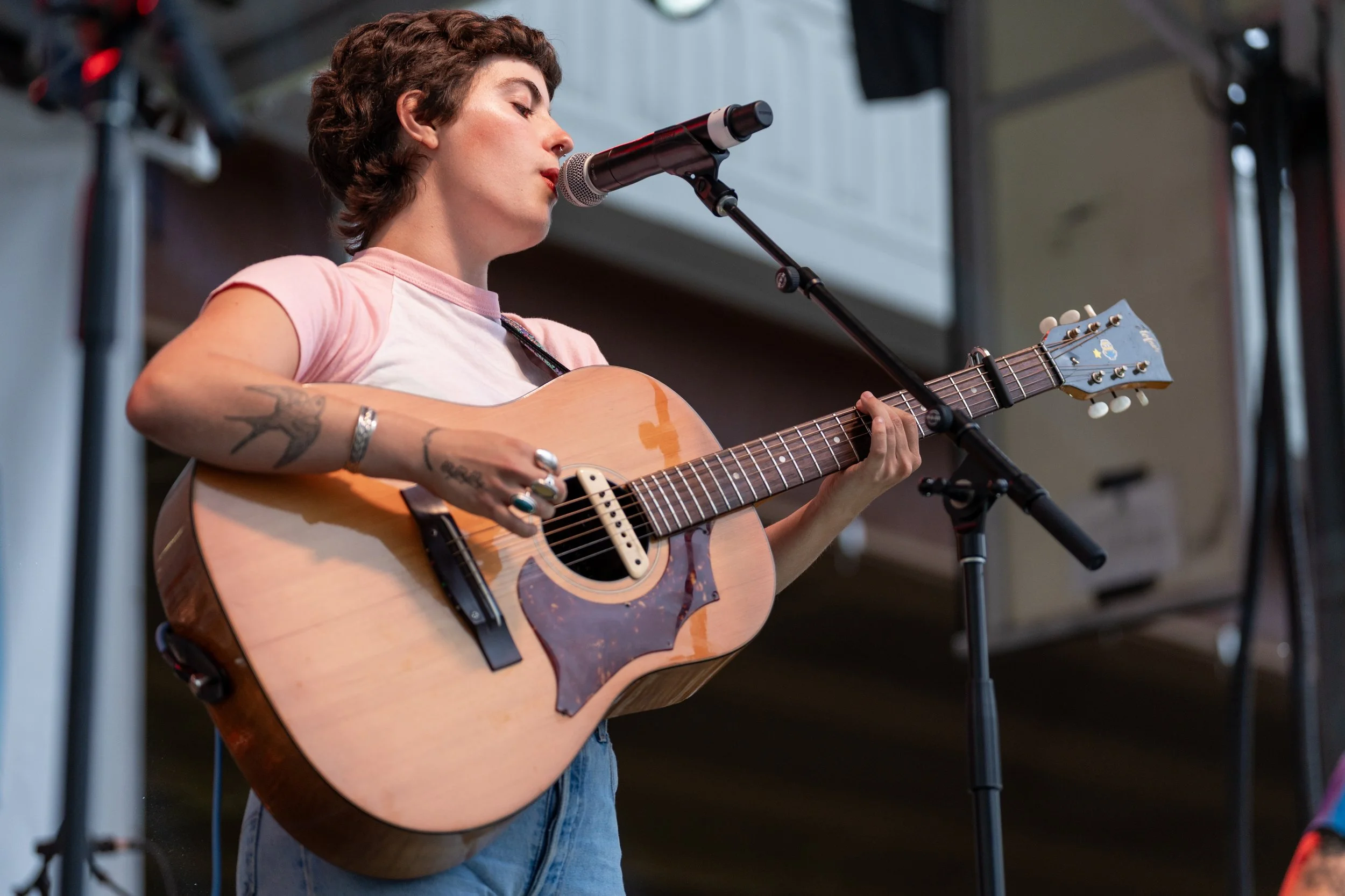 A young woman with short curly hair playing an acoustic guitar and singing into a microphone at an indoor event.