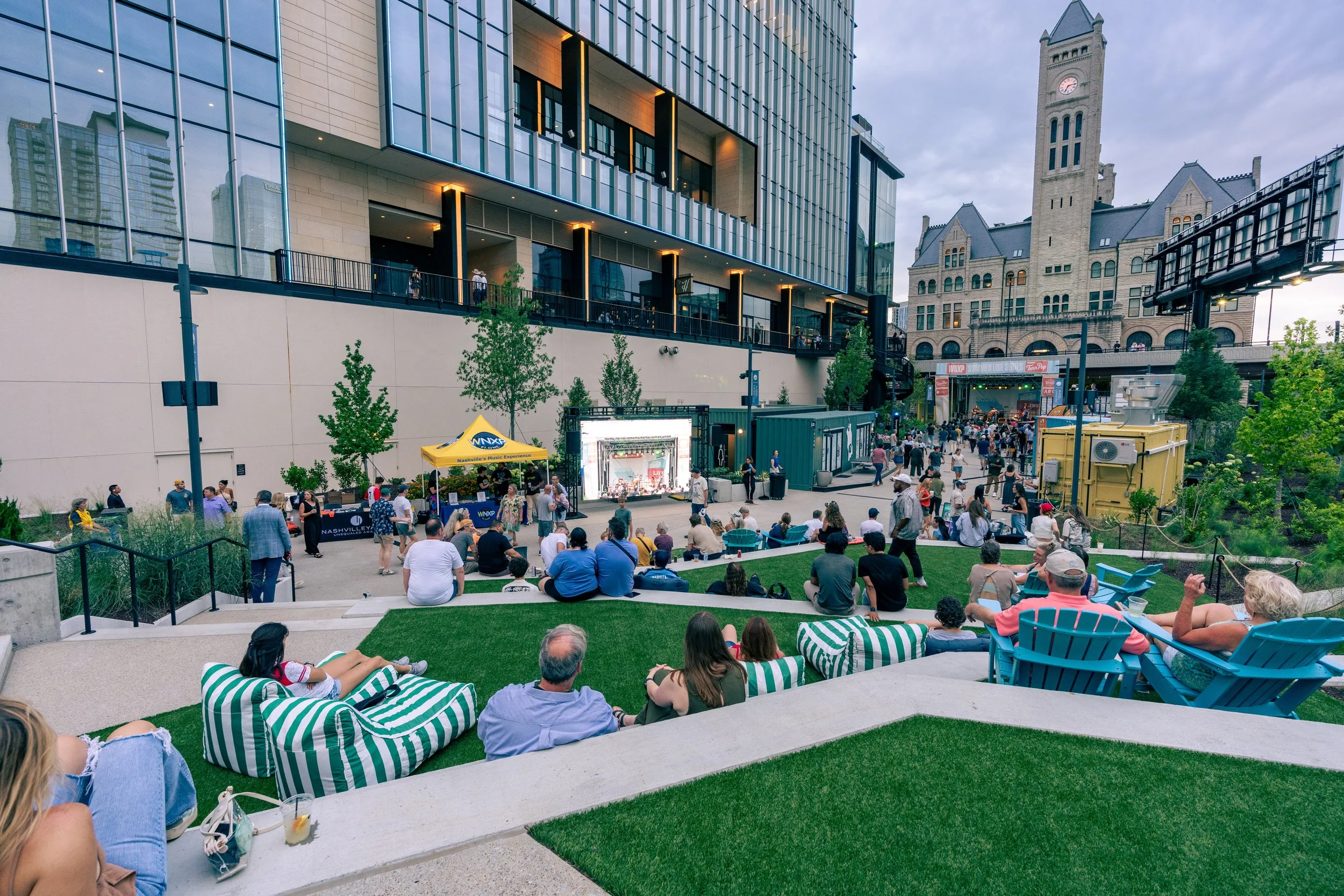 Crowd gathering at an outdoor concert in an urban park setting with modern and historic buildings in the background. People are sitting on green striped bean bag chairs and concrete steps, watching a stage with a band. There is a yellow tent and a large digital screen, with trees and pathways around.