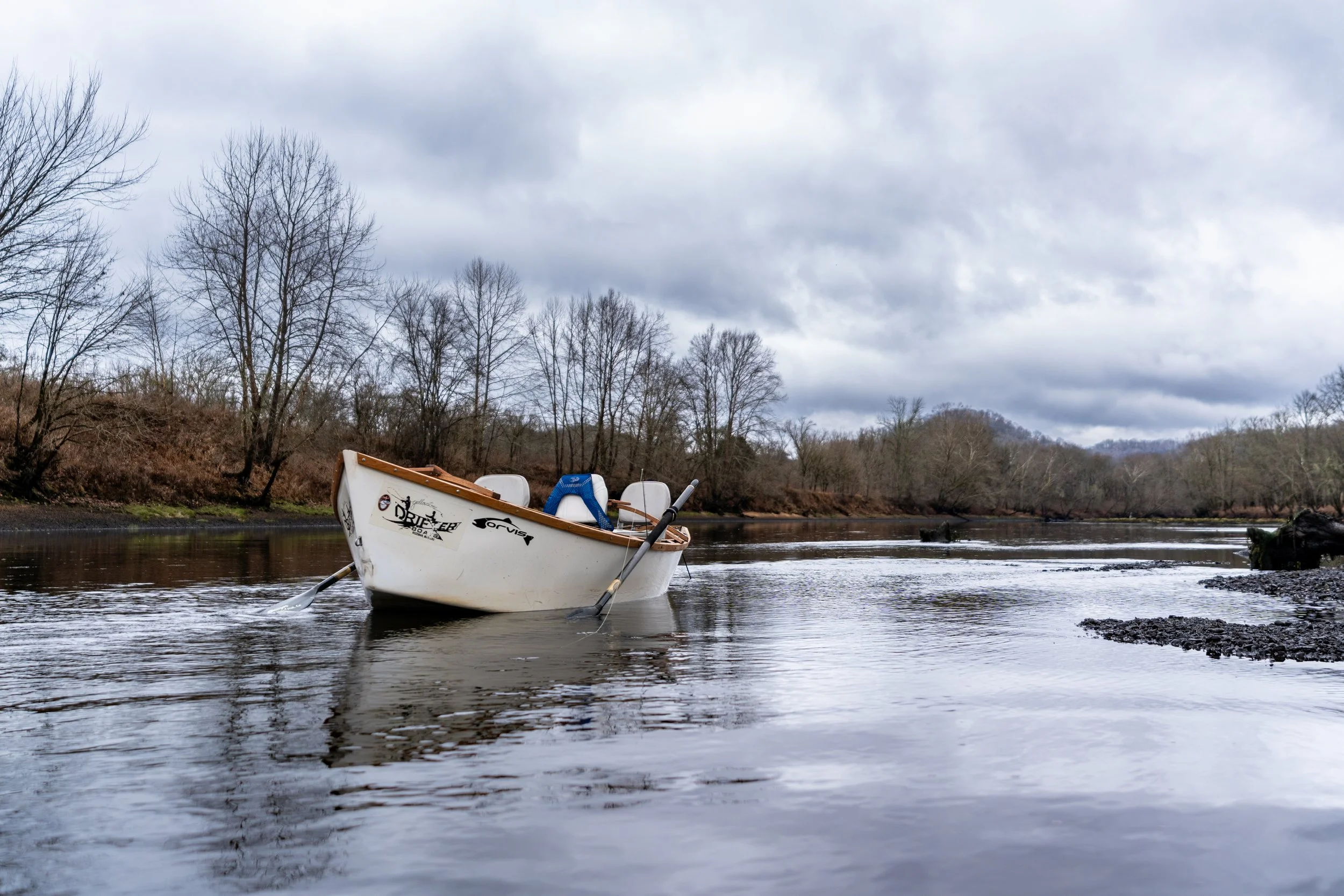 A white boat named 'Dreyer' floats on a calm river with a small dog paddling nearby. The boat is surrounded by leafless trees on the riverbank under a cloudy sky.