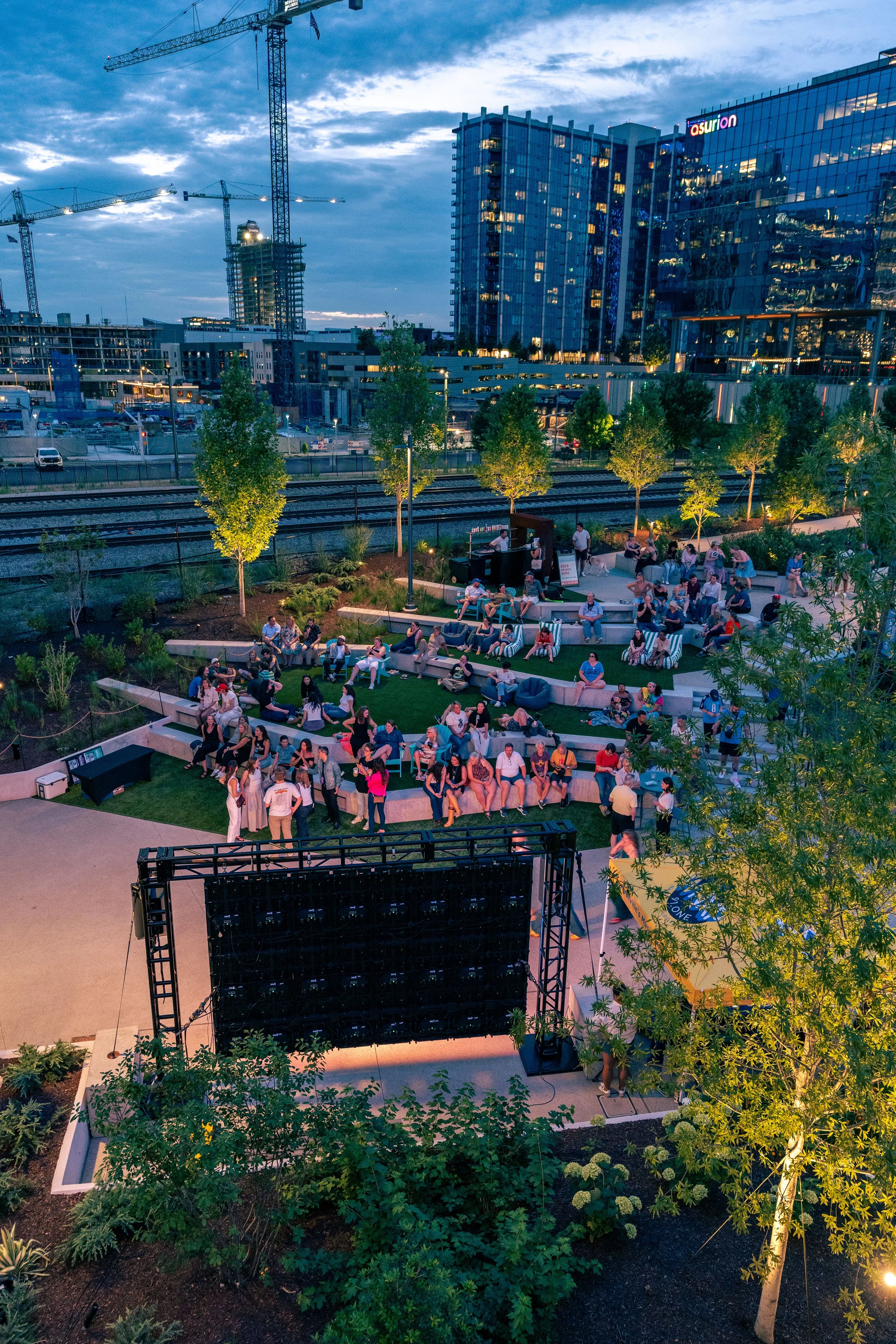 An outdoor music venue with a large empty stage and audience seating area with people gathered, surrounded by trees and city buildings with construction cranes in the background during dusk.