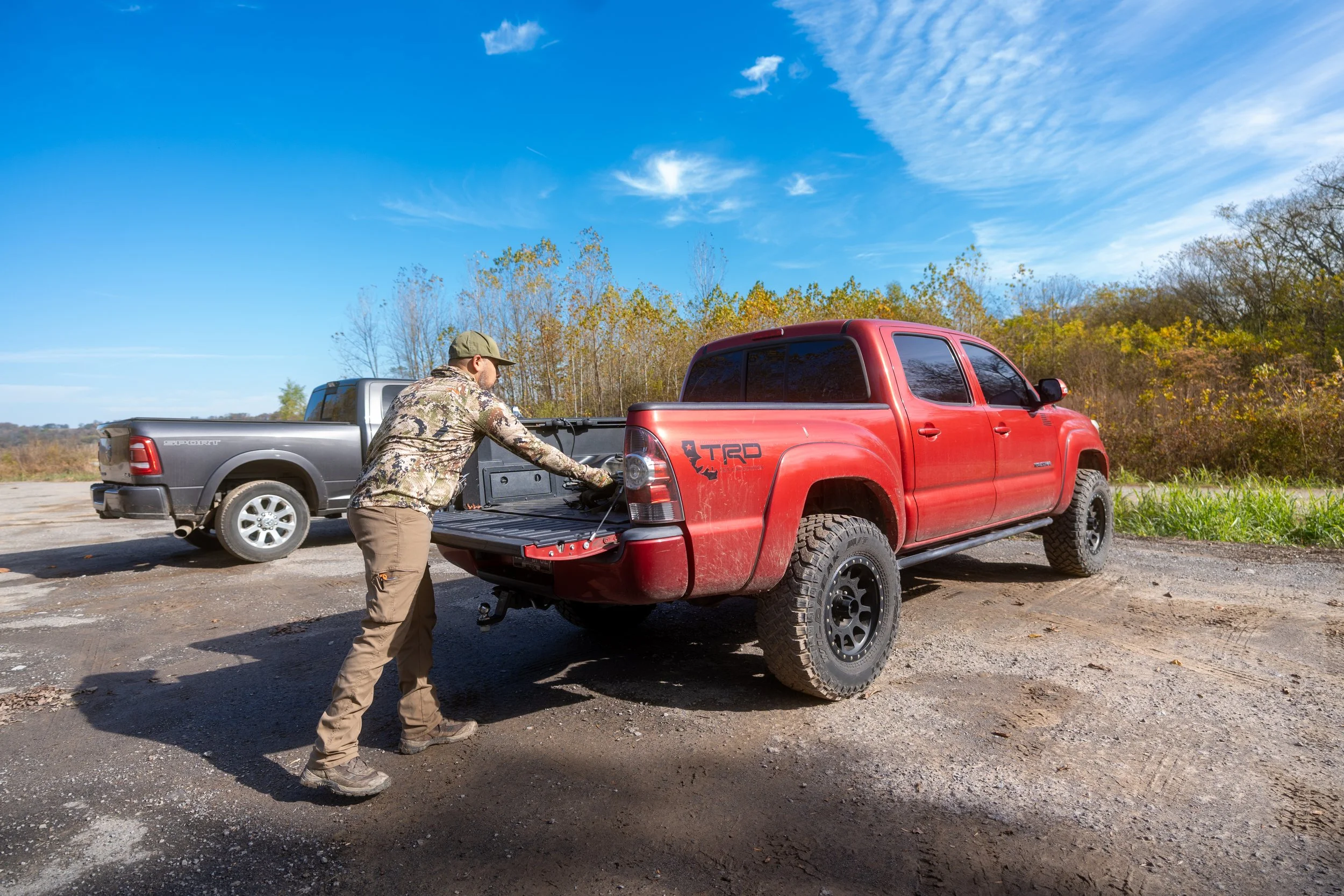 A man wearing camouflage jacket and cap is working on the tailgate of a red pickup truck in an outdoor setting with trees and blue sky in the background.