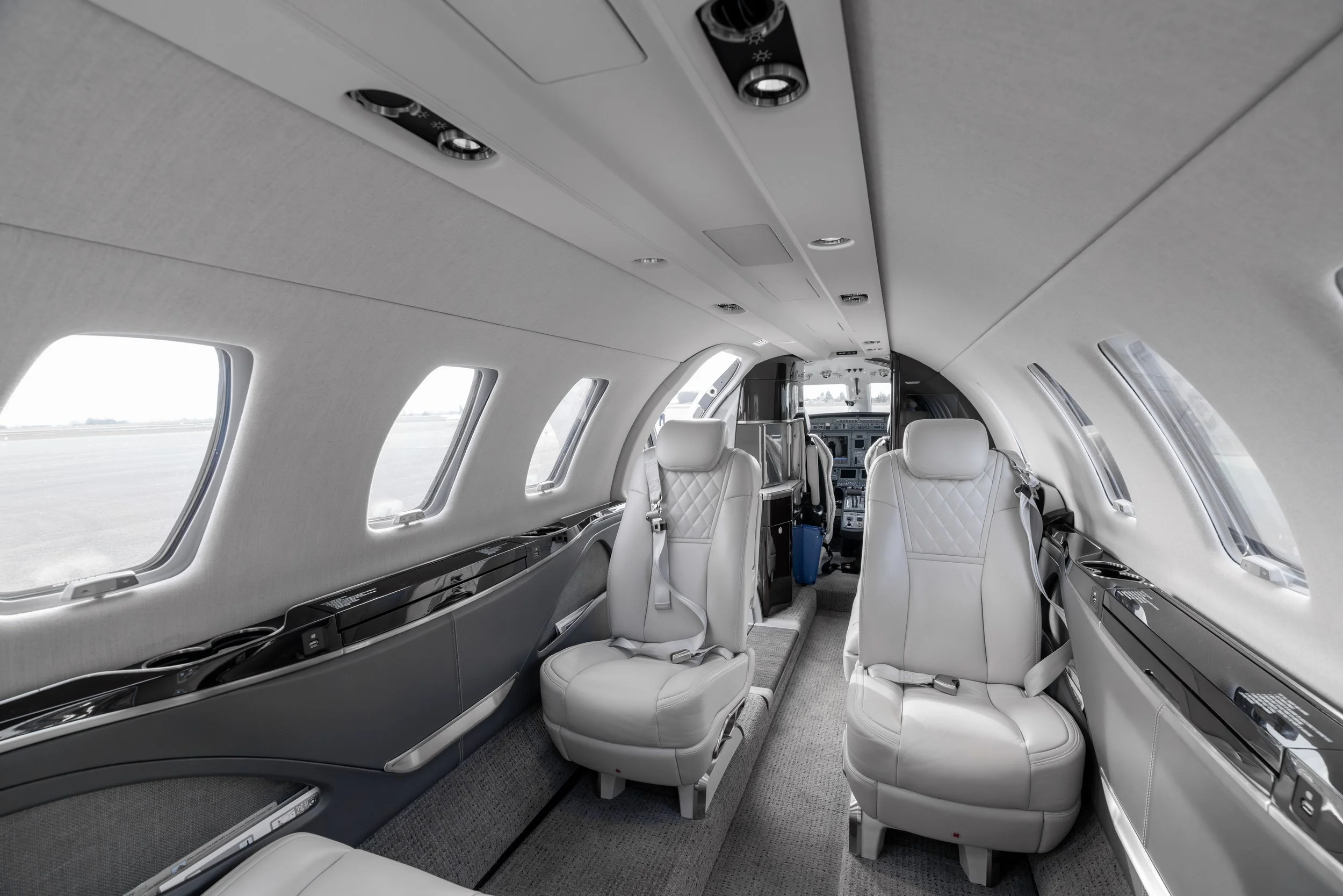 Interior of a private jet with white leather seats, windows, and overhead lighting, showing the cockpit area in the front.