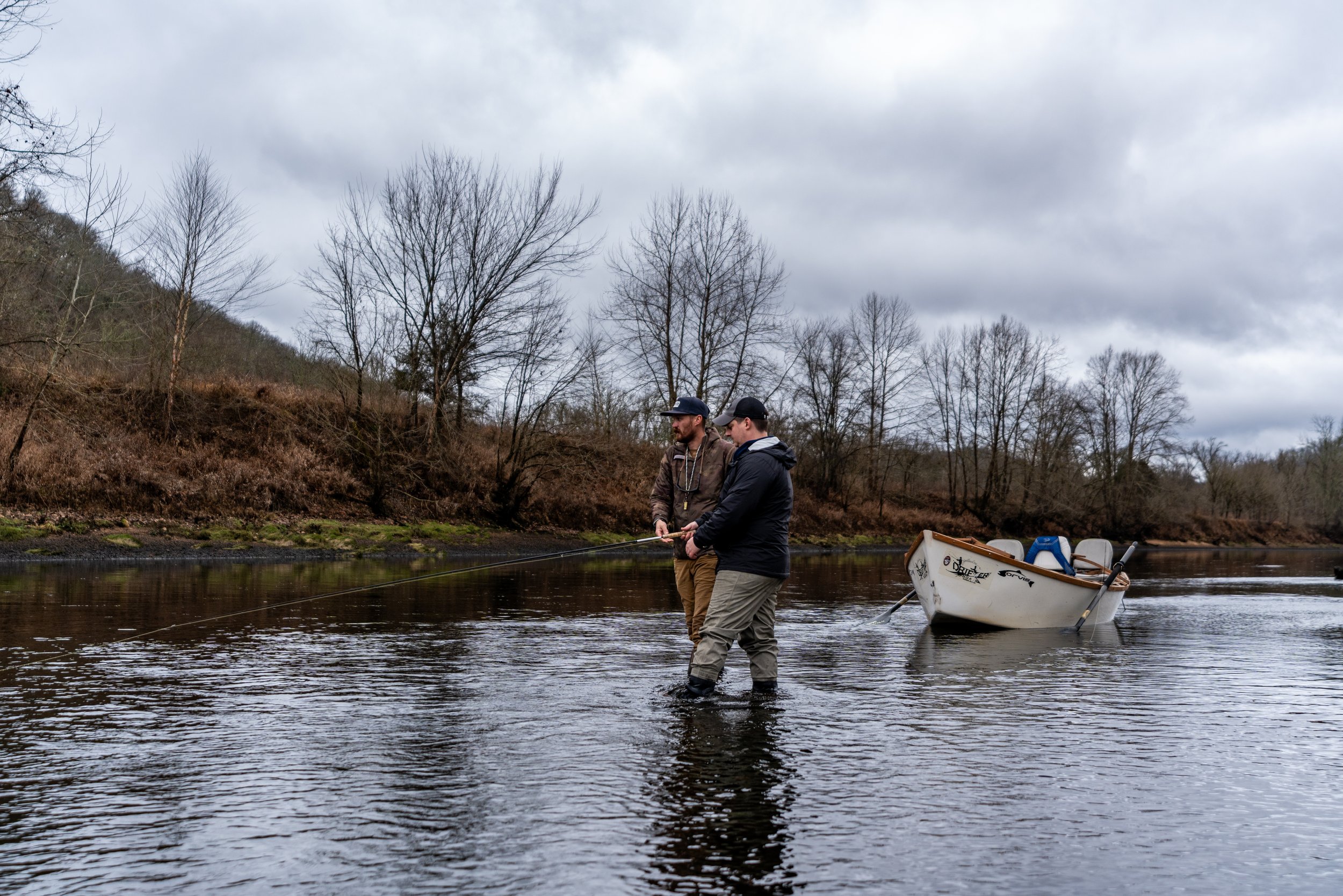 Two men are fishing in a river with a boat near the shore. They are dressed in jackets and hats, standing in the water. In the background, there are leafless trees on a hillside under a cloudy sky.