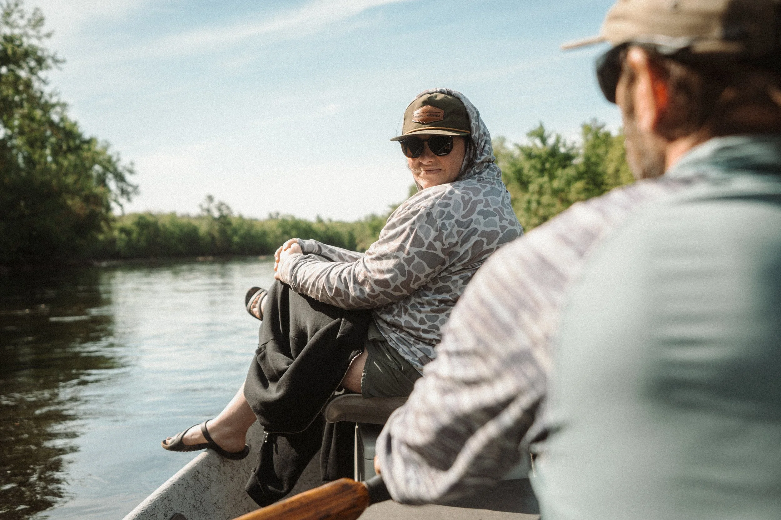 Two people sitting in a canoe on a river, surrounded by greenery, enjoying a sunny day.