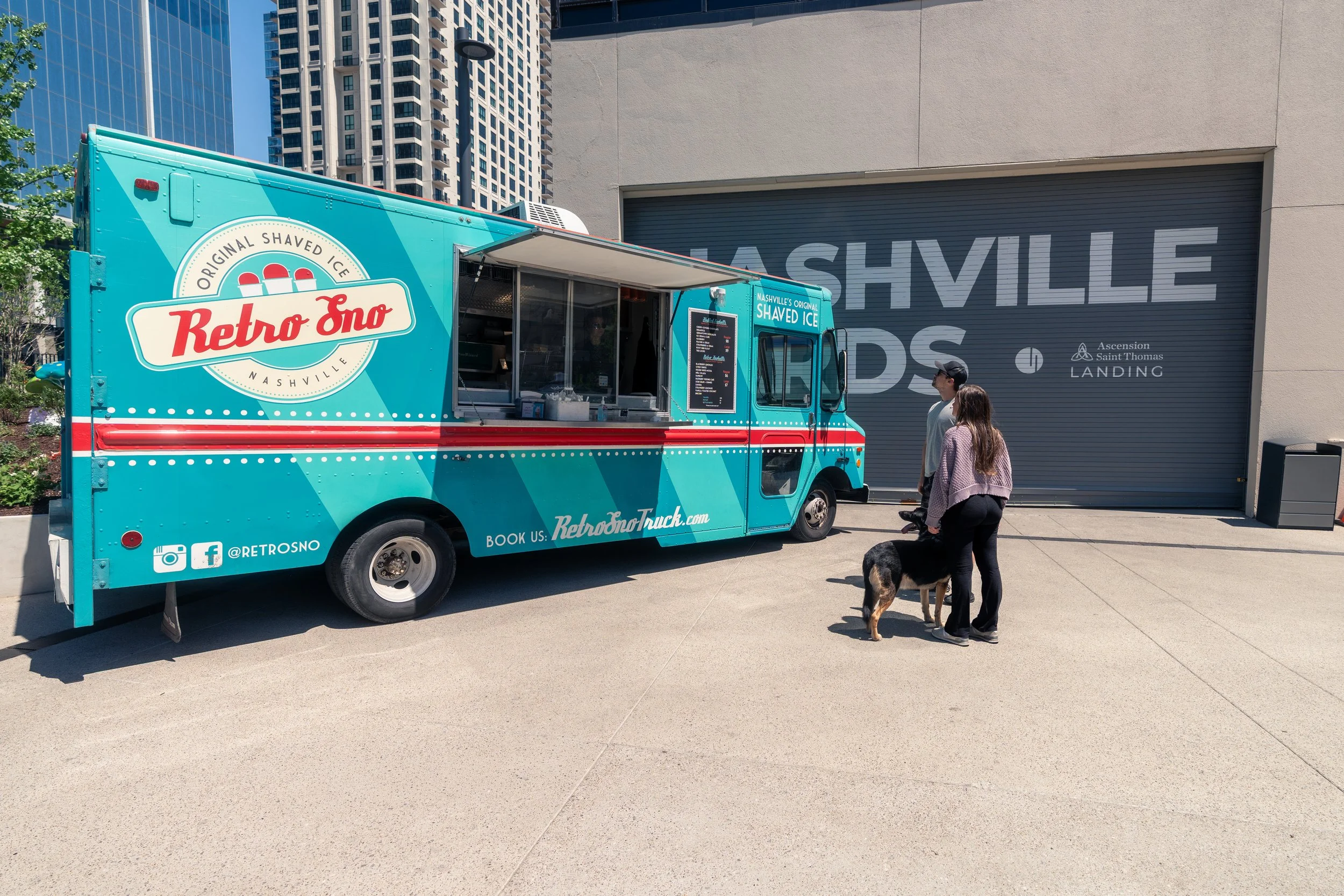 A bright blue Retro Uno shaved ice truck is parked on a sidewalk near modern high-rise buildings. Two women are standing in front of the truck, one with a dog, engaging in conversation. The truck has social media icons and a website link displayed on