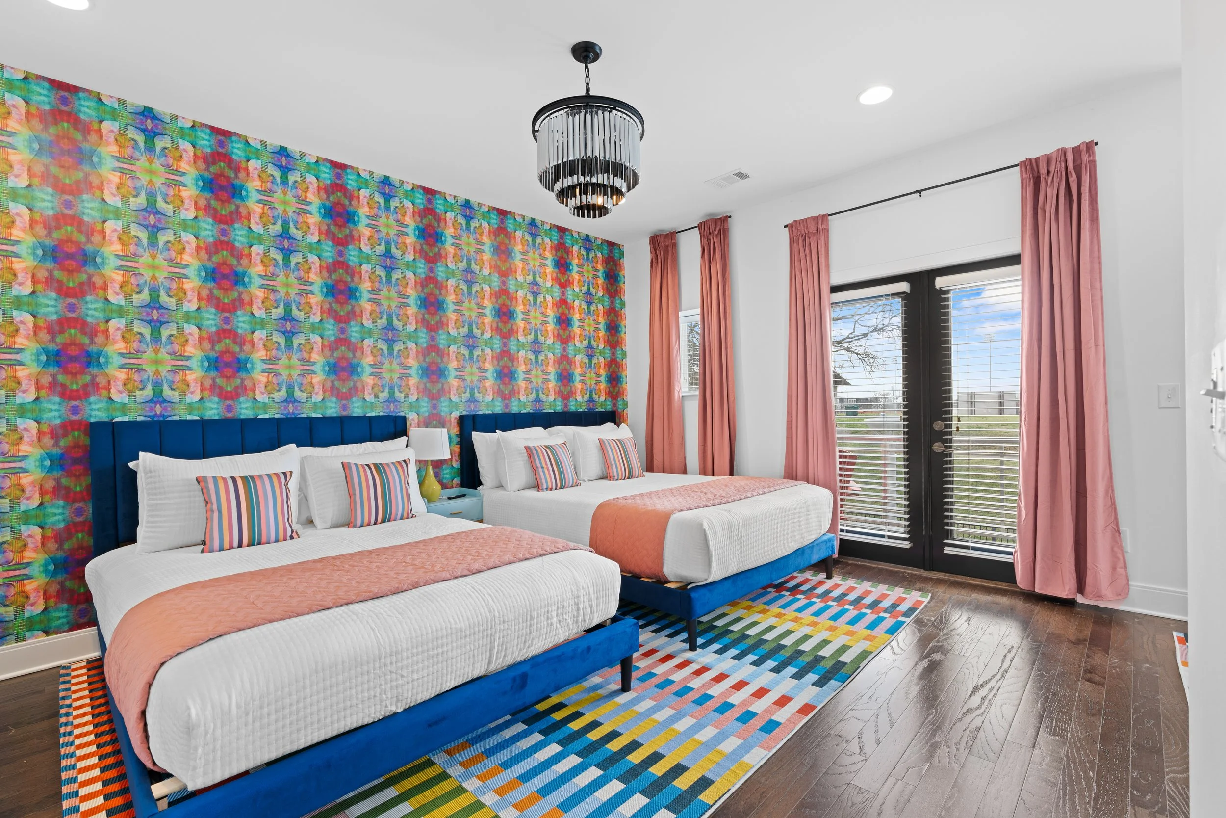 A brightly decorated bedroom with two beds featuring white linens and colorful striped pillows, a vibrant multicolored wall behind them, pink curtains, a black chandelier, and large windows with blinds showing an outdoor view.