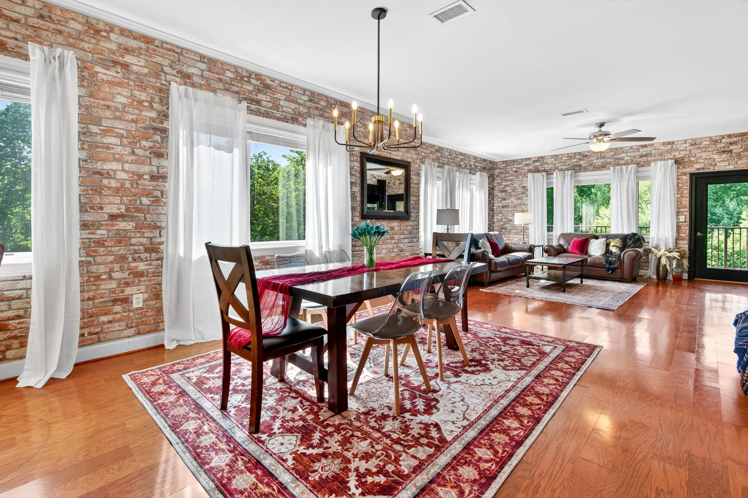 Living room with brick walls, large windows with white curtains, hardwood floors, leather sofas with pillows, a dining table with chairs, a chandelier, table lamps, a mirror, and a door leading outside to a balcony.