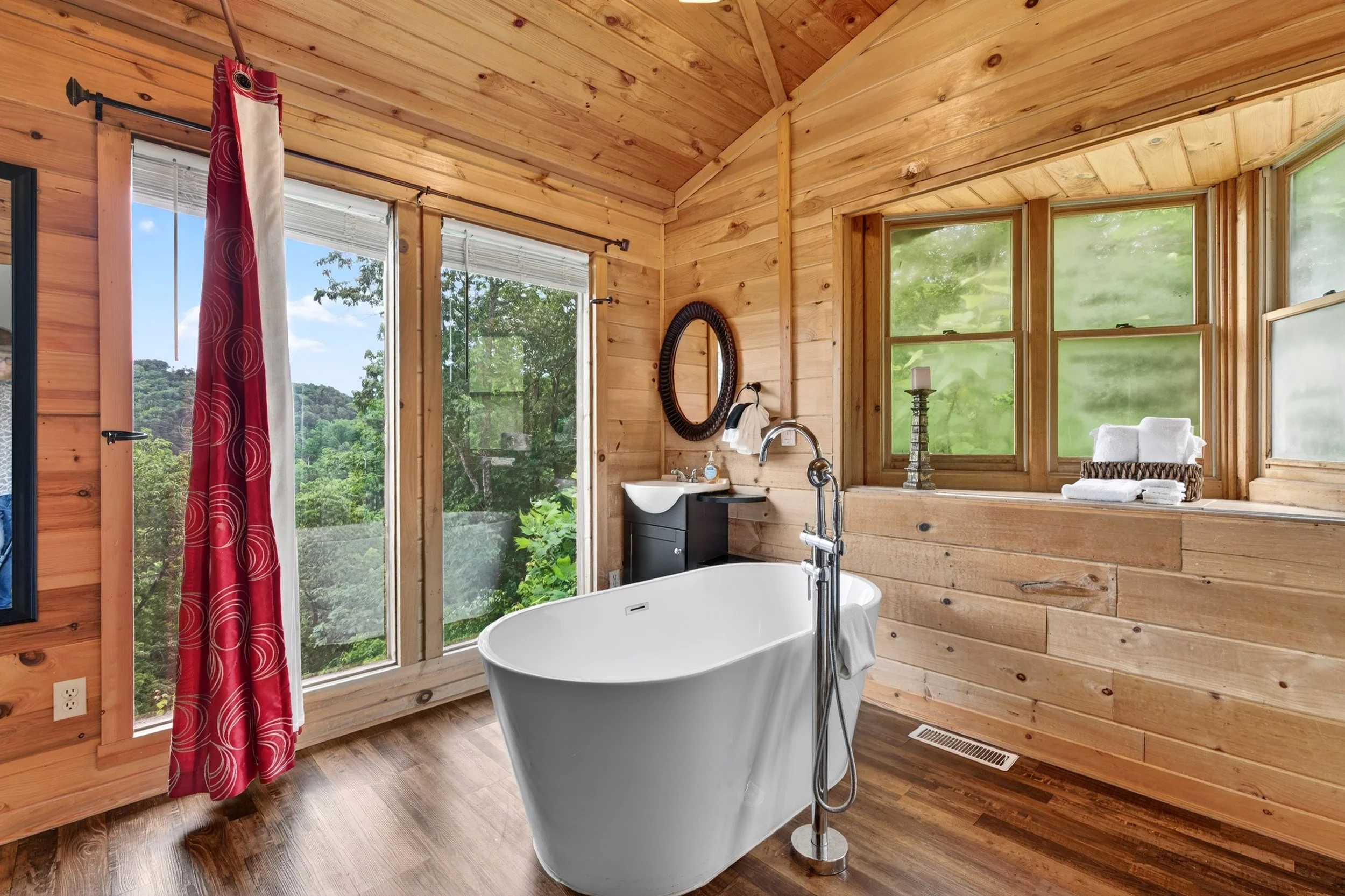A rustic bathroom with wooden walls and ceiling, featuring a large soaking bathtub, a small sink with a mirror, and large windows overlooking a lush, green landscape.