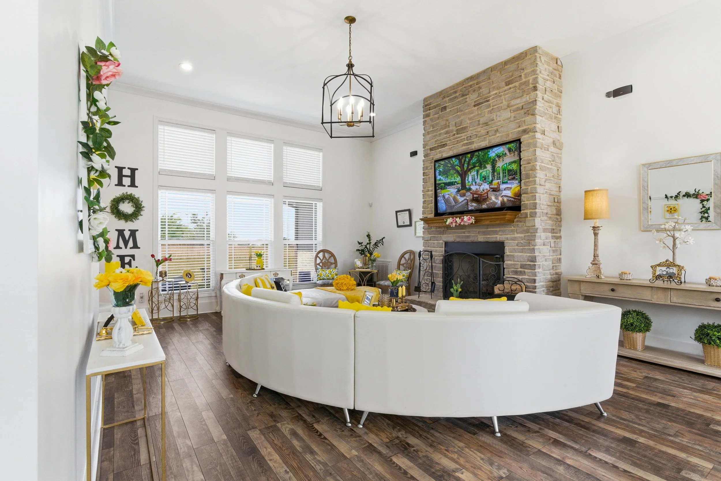 Living room with white sectional sofa, wooden flooring, a mounted TV on a brick fireplace, large windows with white blinds, and decorated with flowers, wreaths, and framed pictures.