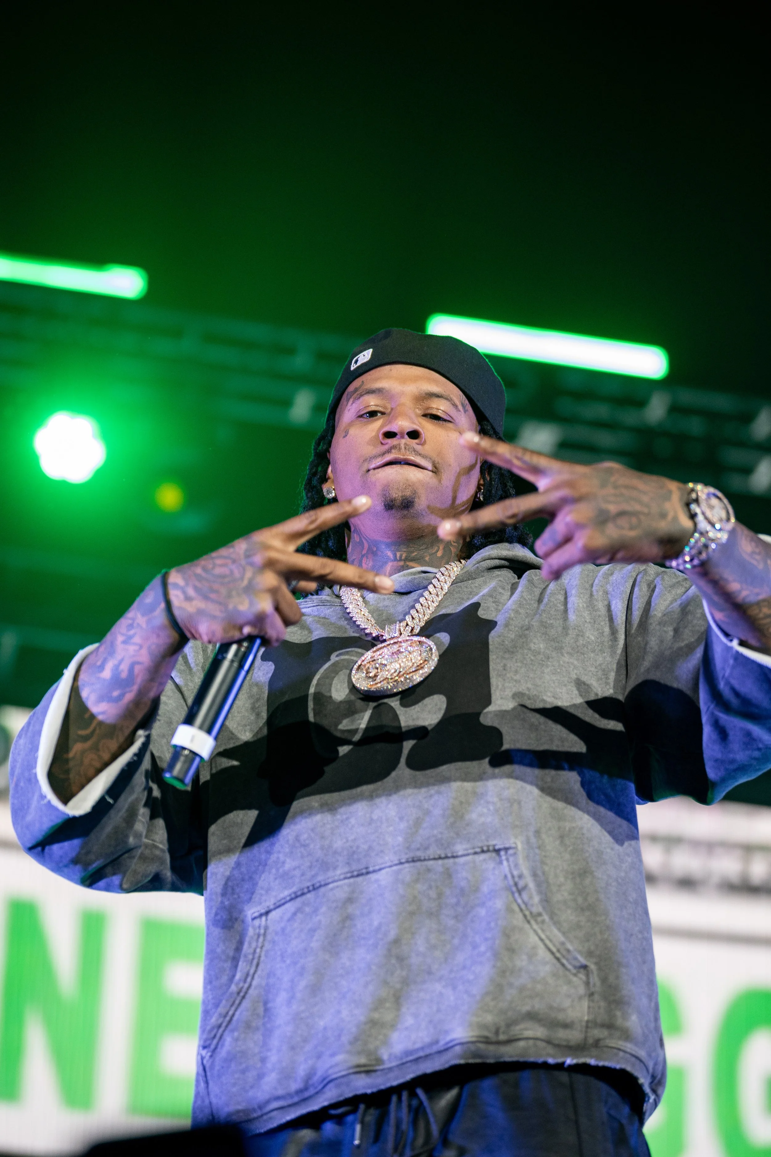 A man with tattoos, wearing a black cap and a gray camouflage hoodie, making a hand gesture with both hands, holding a microphone in his right hand. Bright green stage lights are in the background.