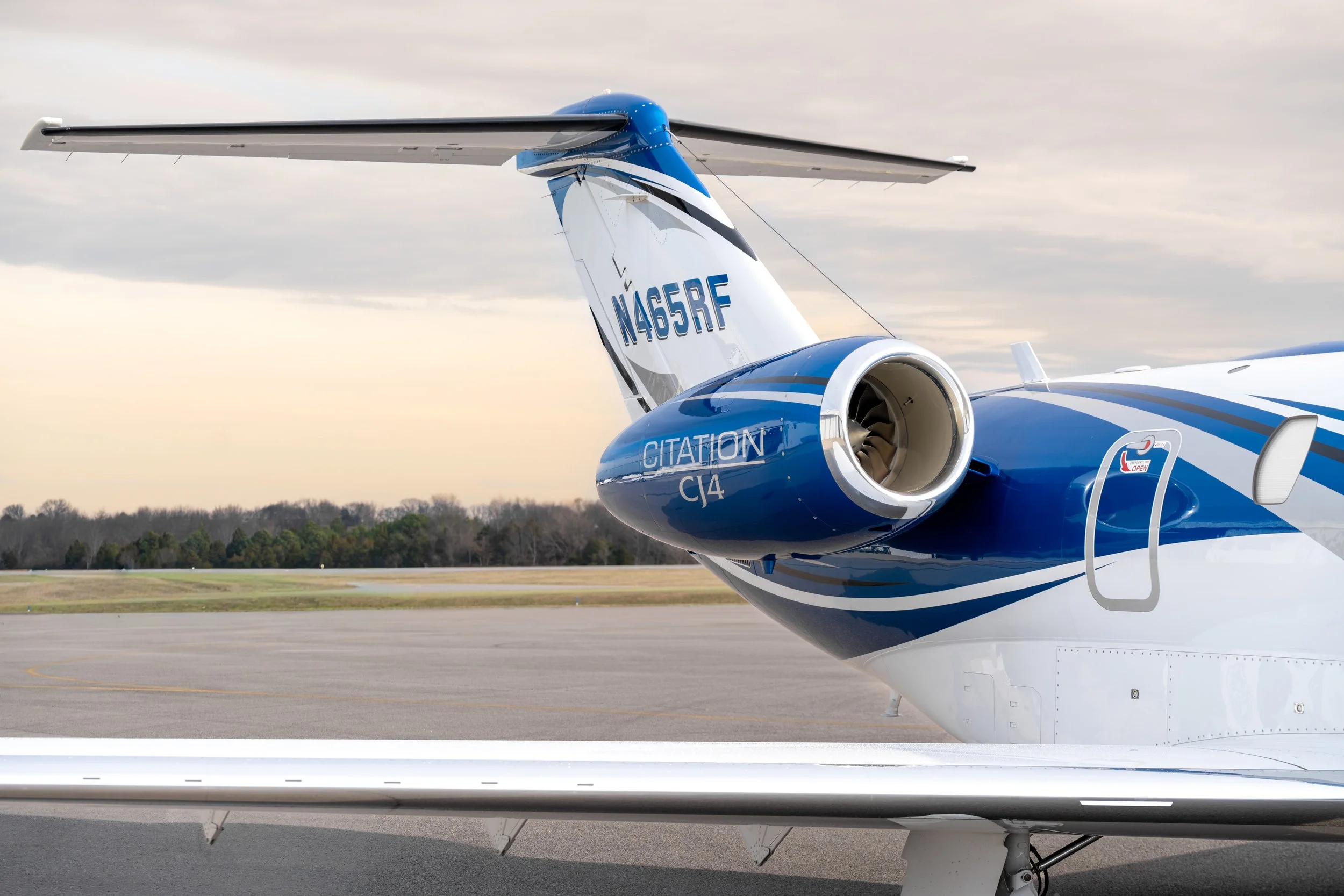 Close-up of a modern blue and white private jet aircraft on the tarmac, with a visible tail, jet engine, and part of the wing showing. Overcast sky in the background.