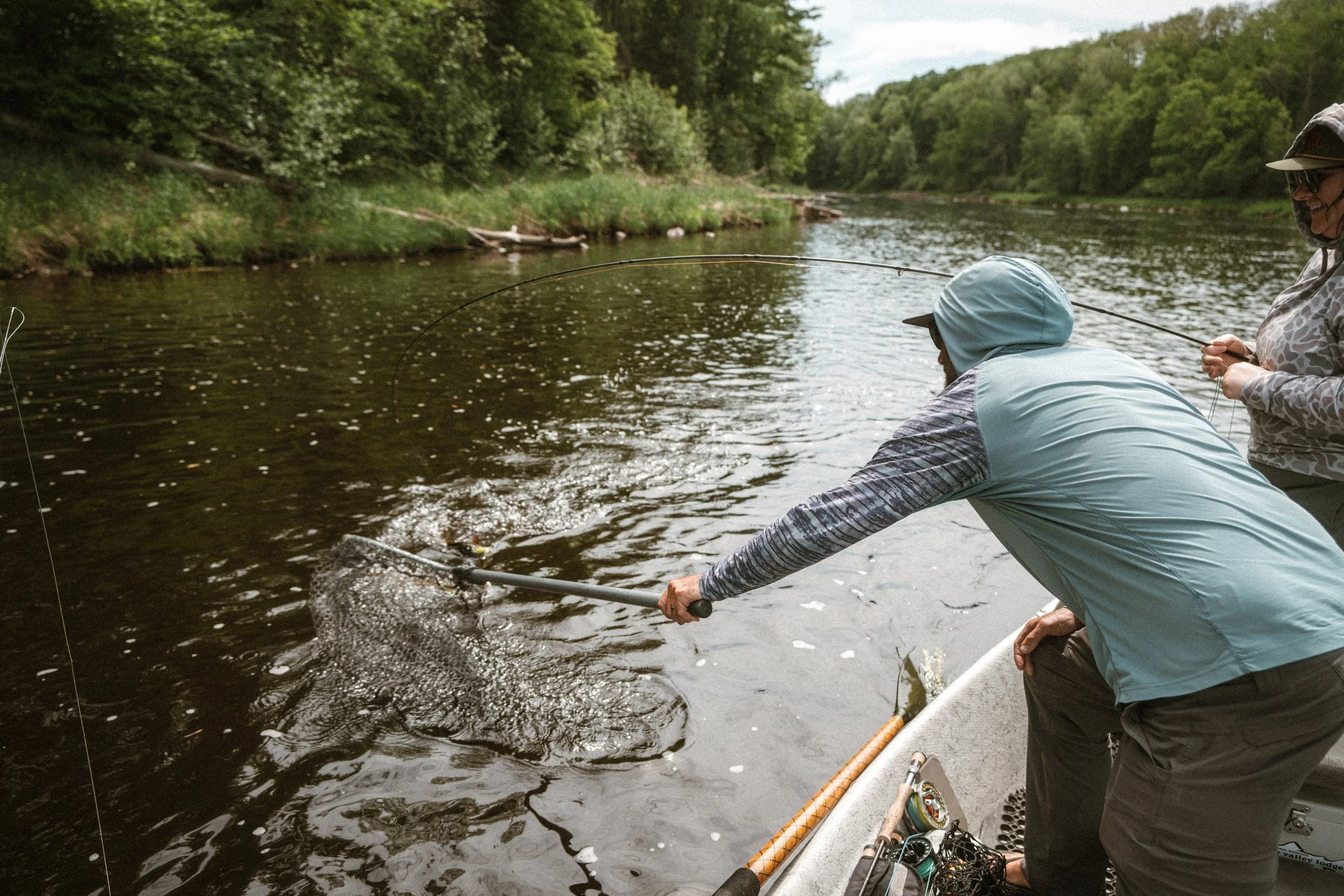 Two people fishing in a river surrounded by lush green trees, one person is bending over holding a fishing net with a fish in it, and the other is standing, smiling, holding a fishing rod.