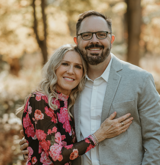 A smiling couple outdoors, woman in a floral dress and man in a light gray suit, embracing each other.