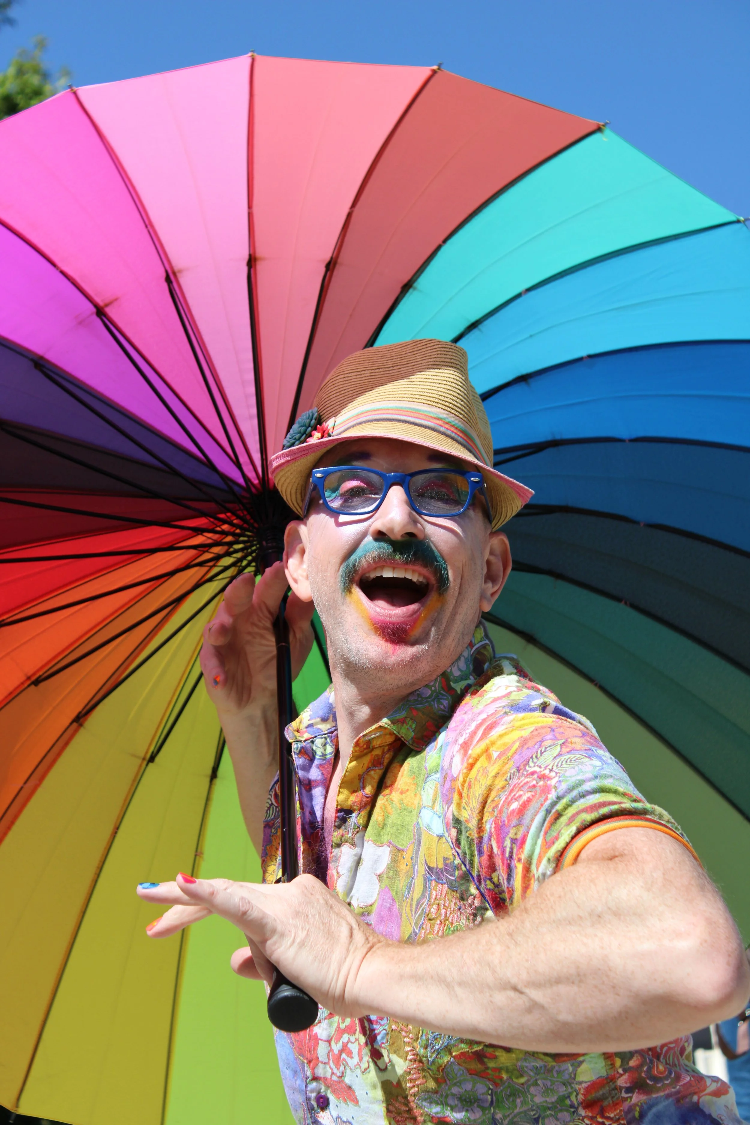 A person with rainbow-painted lips and sunglasses taking a selfie outdoors under a large rainbow-colored umbrella, wearing a floral shirt and a straw hat.