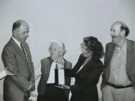 Four people standing together, with one woman presenting an award or plaque to an elderly woman, in a formal setting.