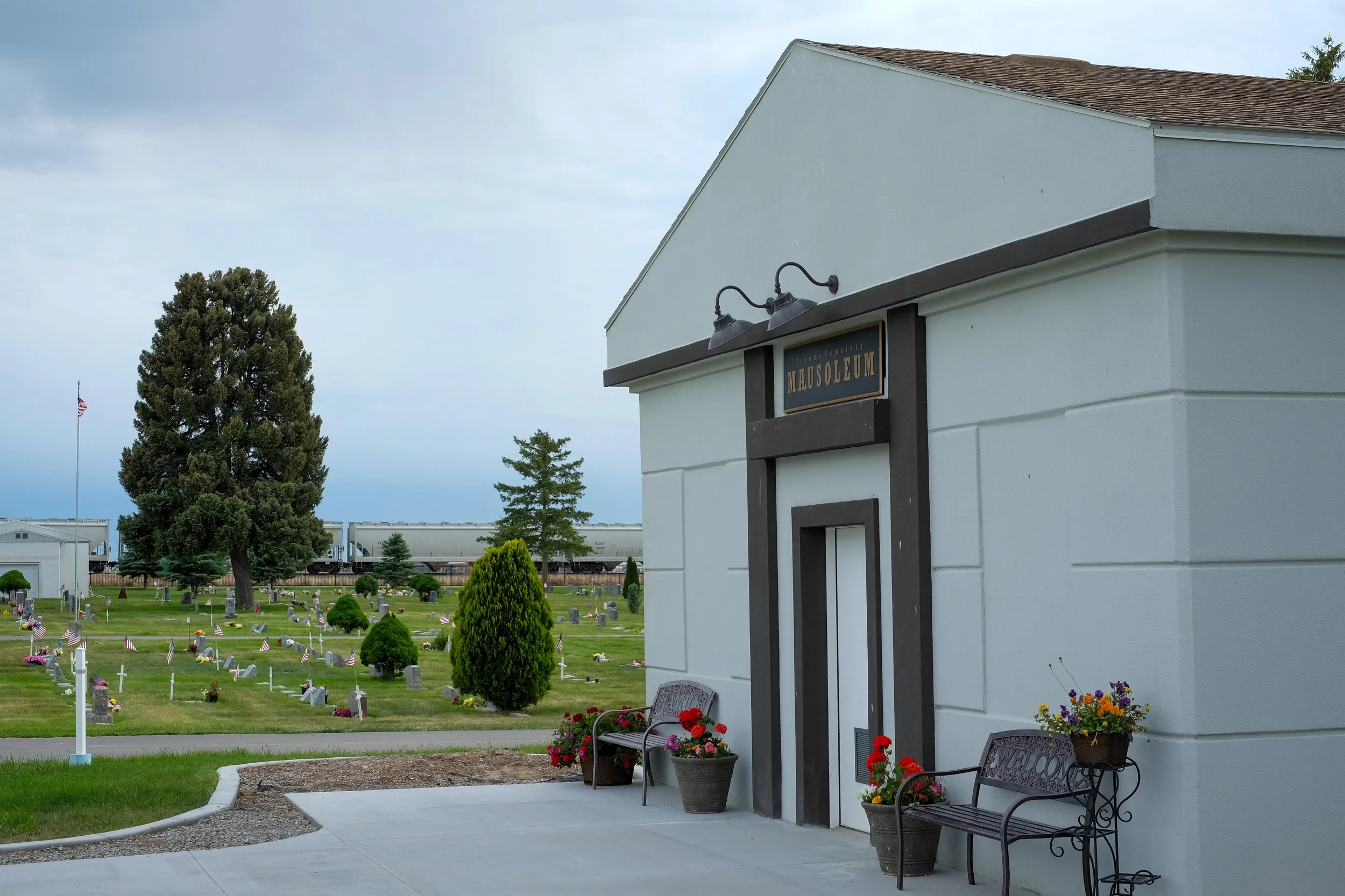 White mausoleum building with black trim and an approaching graveyard with headstones and American flags, surrounded by trees and benches.