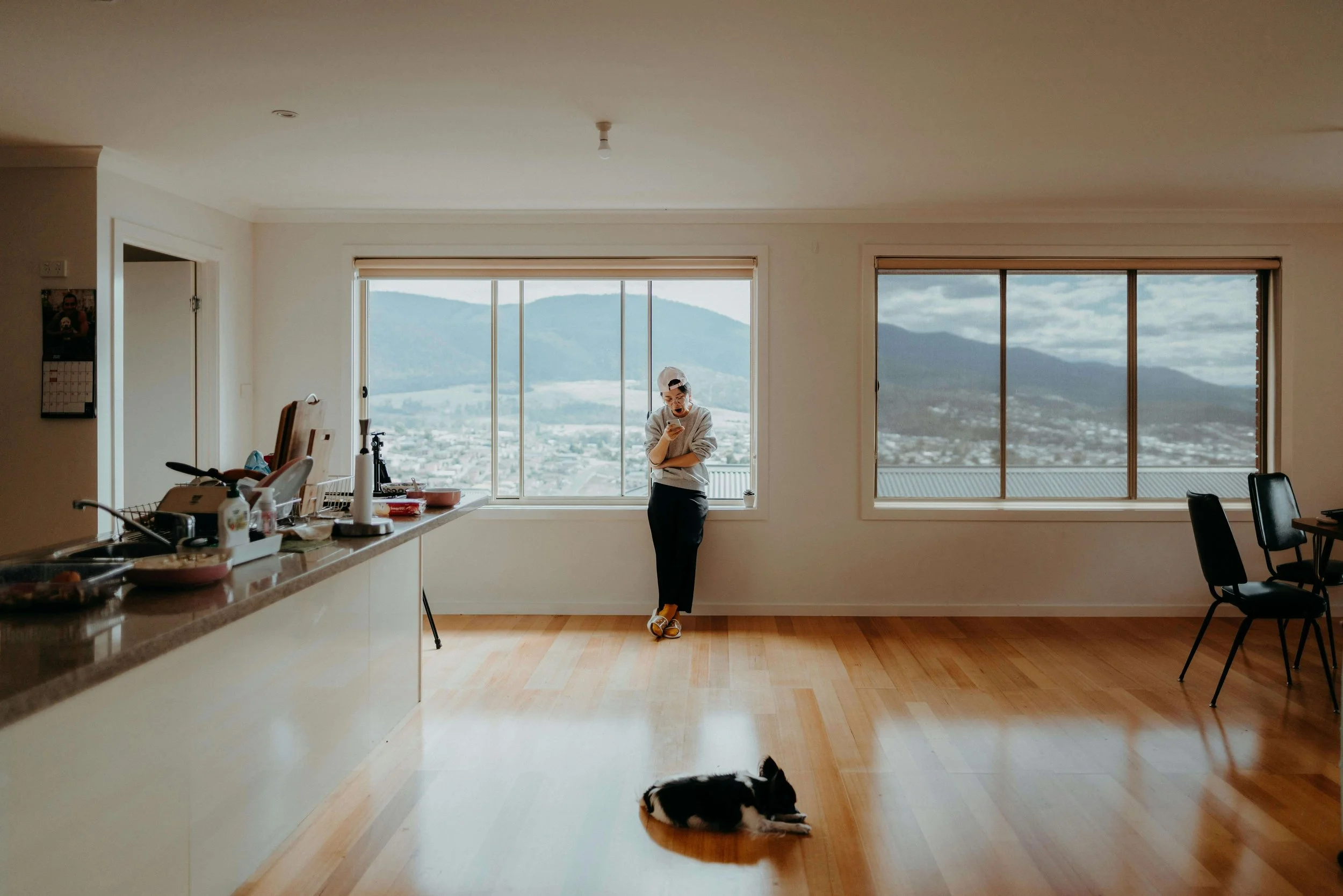 A woman in a gray hoodie and cap standing in a bright living room, looking at her phone, with a black and white puppy lying on the wooden floor nearby and mountains visible through the windows.