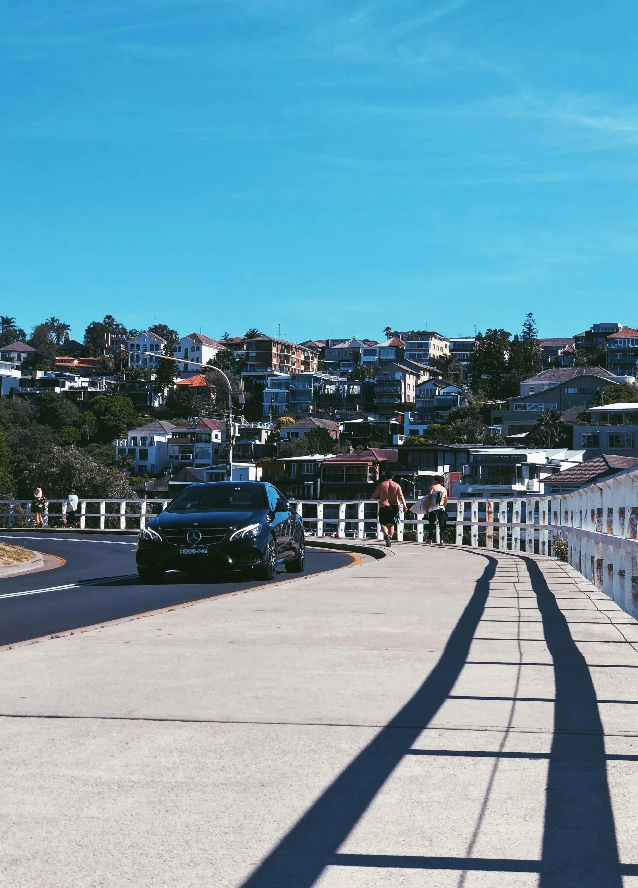 A black Mercedes-Benz car driving along a curved road with a white turnpike railing, a man and a woman walking nearby, low hill with residential houses and greenery, clear blue sky.