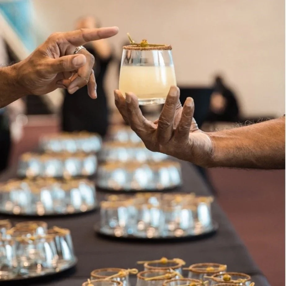 Two people exchanging a cocktail with a cinnamon stick garnish, with multiple similar drinks on a table covered with a black tablecloth in the background.