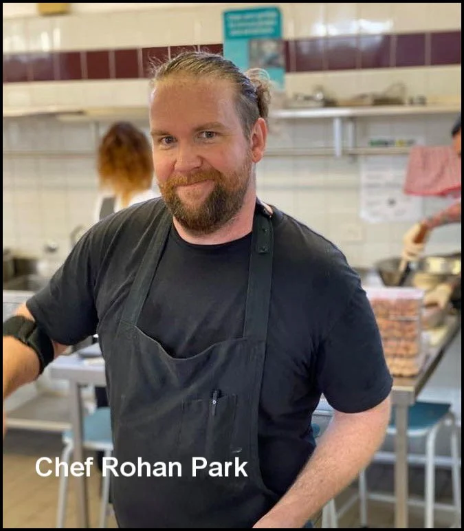 Chef Rohan Park wearing a black apron and t-shirt, smiling in a kitchen setting with other staff in the background.