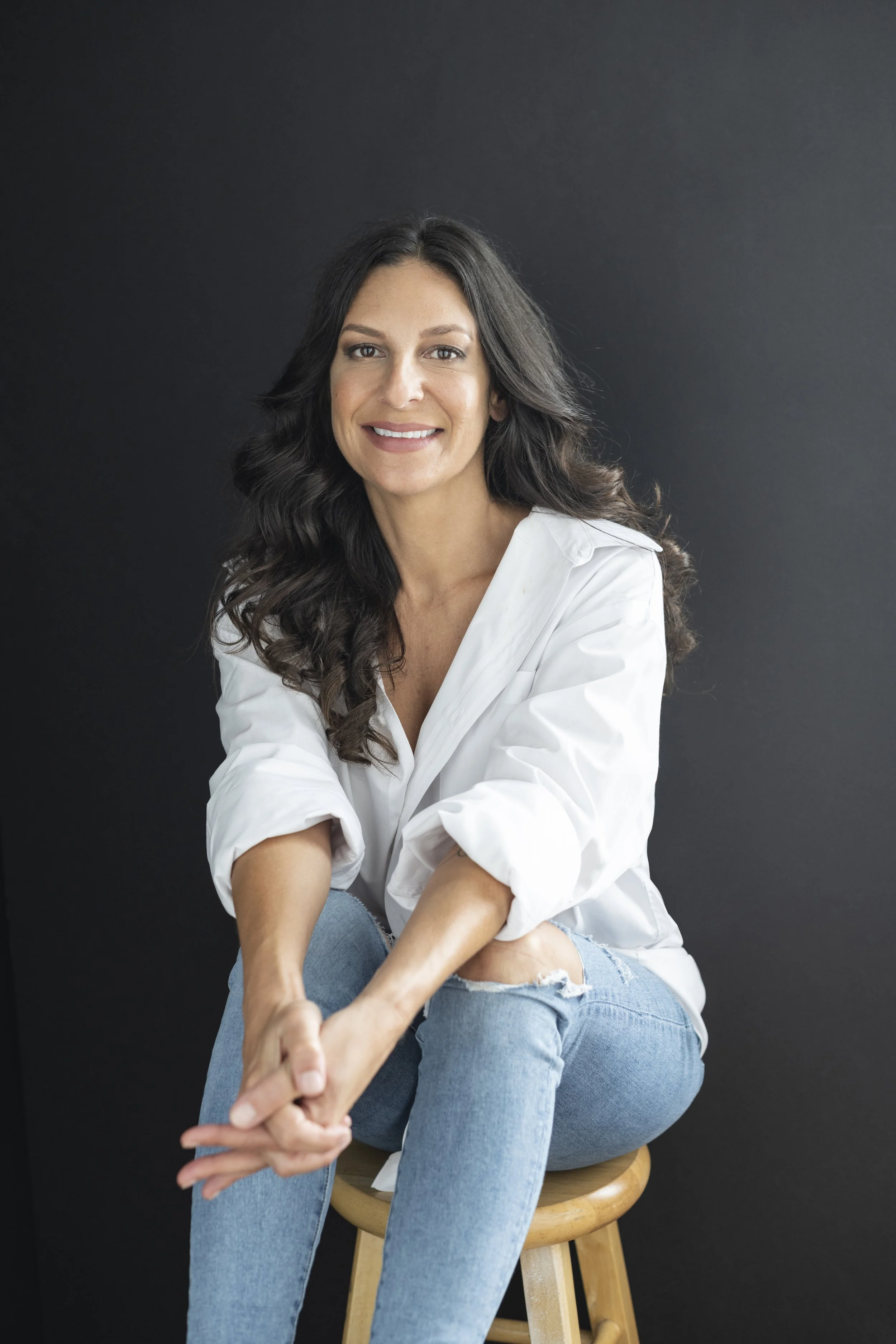A woman with long dark hair sitting on a wooden stool against a black background, wearing a white shirt and ripped jeans, smiling at the camera.