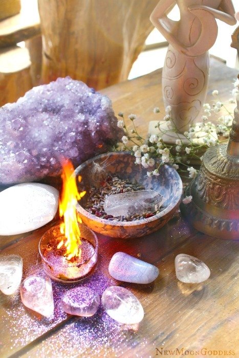 A lit flame on a small copper bowl filled with crystals, set on a wooden table surrounded by various stones, a purple geode, a white ceramic egg, a figurine, and small white flowers.