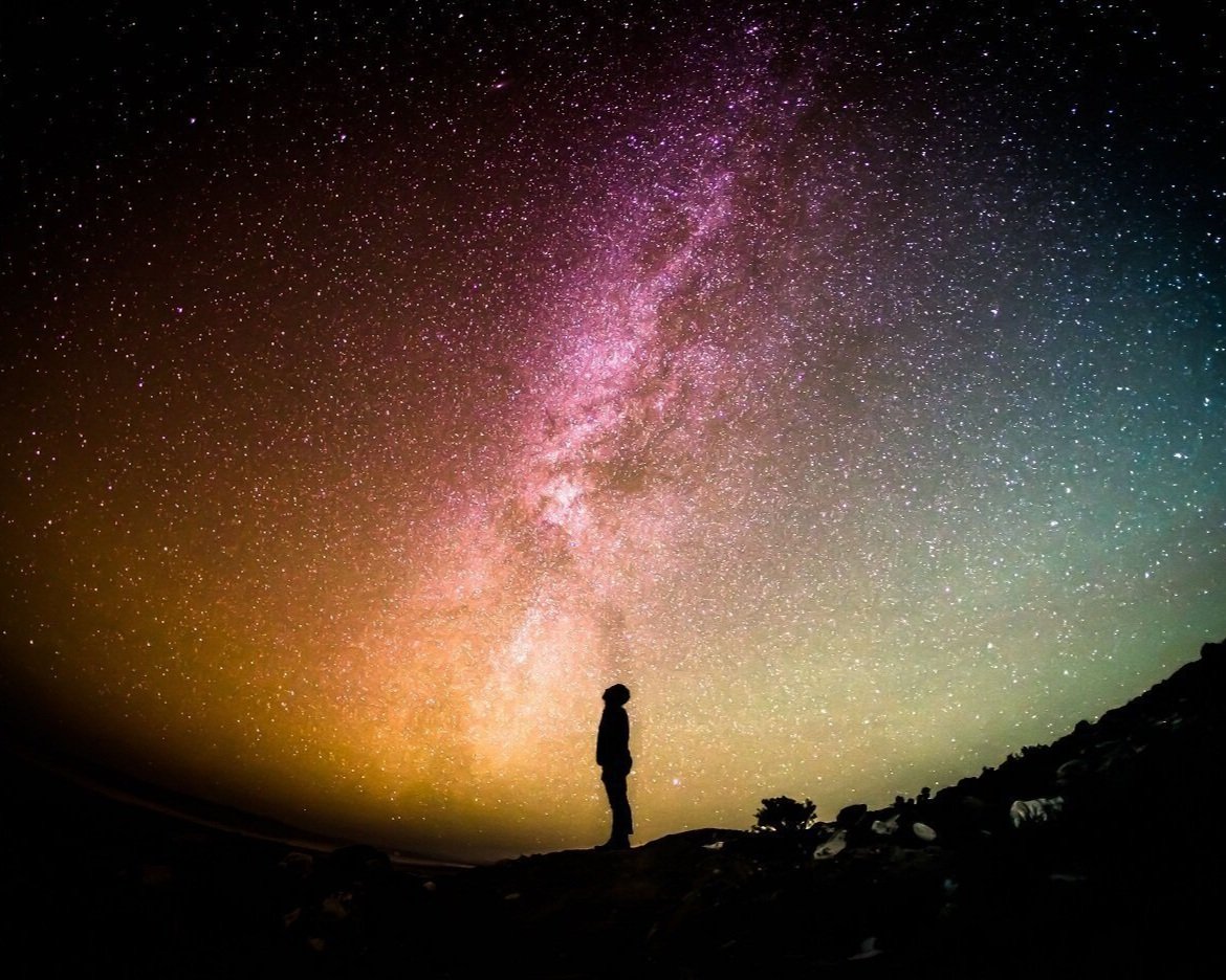 A person standing on a hill at night gazing at a vibrant, star-filled sky with the Milky Way galaxy visible.
