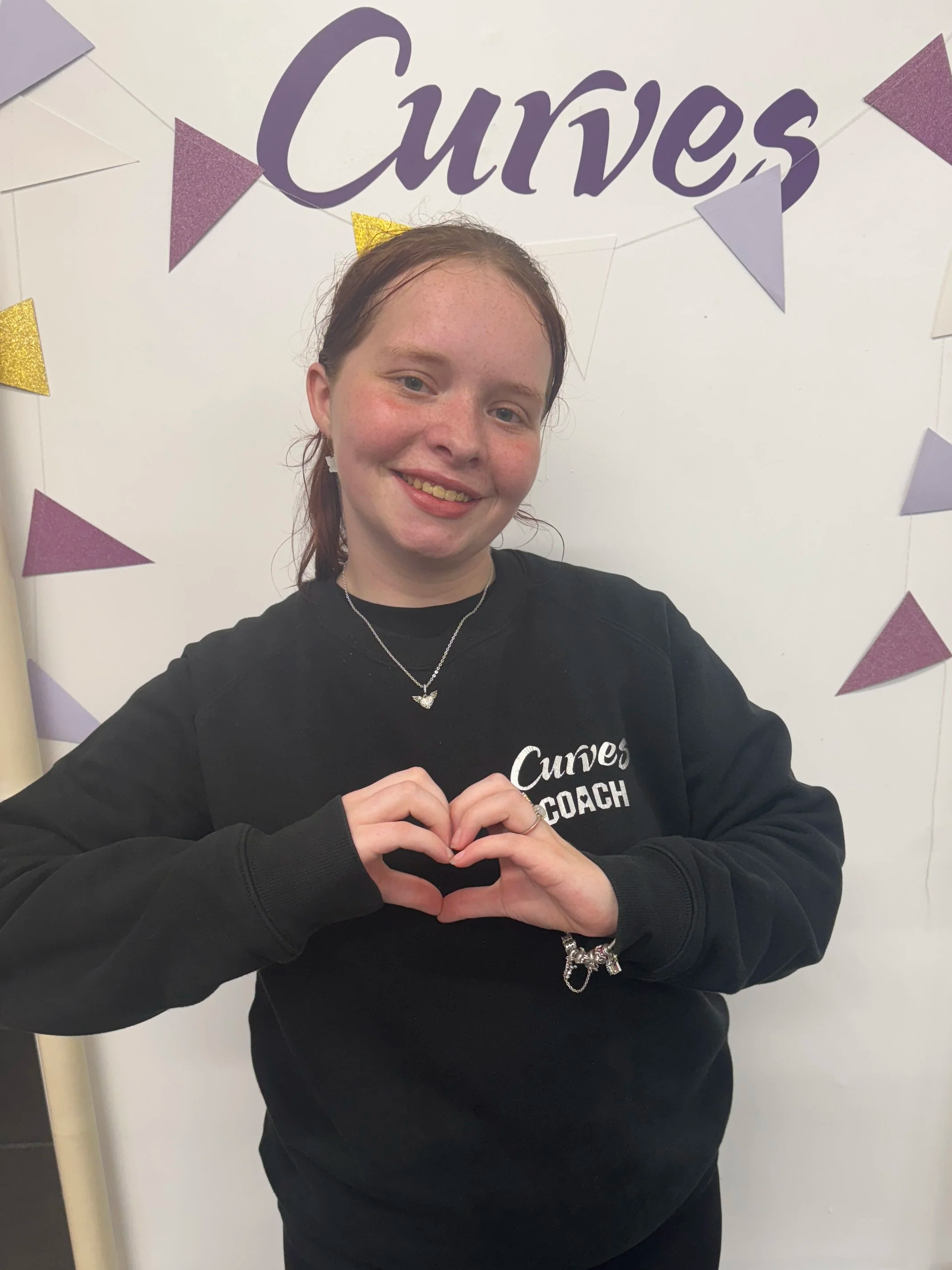 Young woman wearing a Curves Coach sweatshirt smiling beneath the Curves logo, she is making a heart sign with her fingers