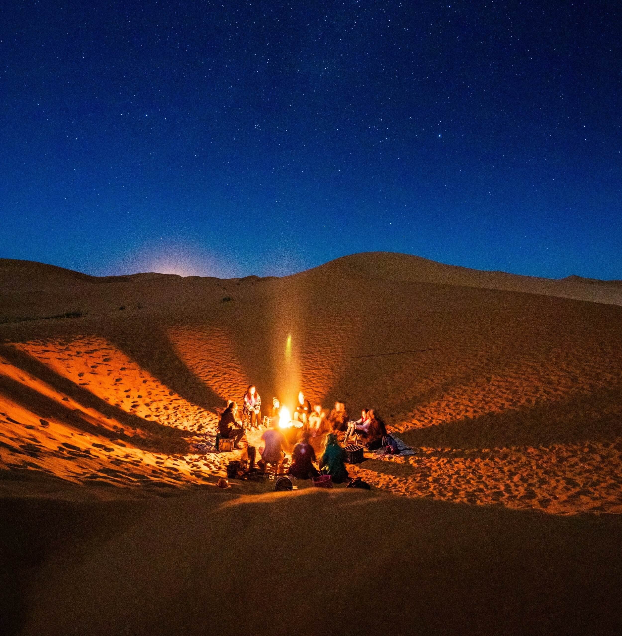 A group of people sitting around a campfire in a desert at night, with a starry sky and mountains in the background.