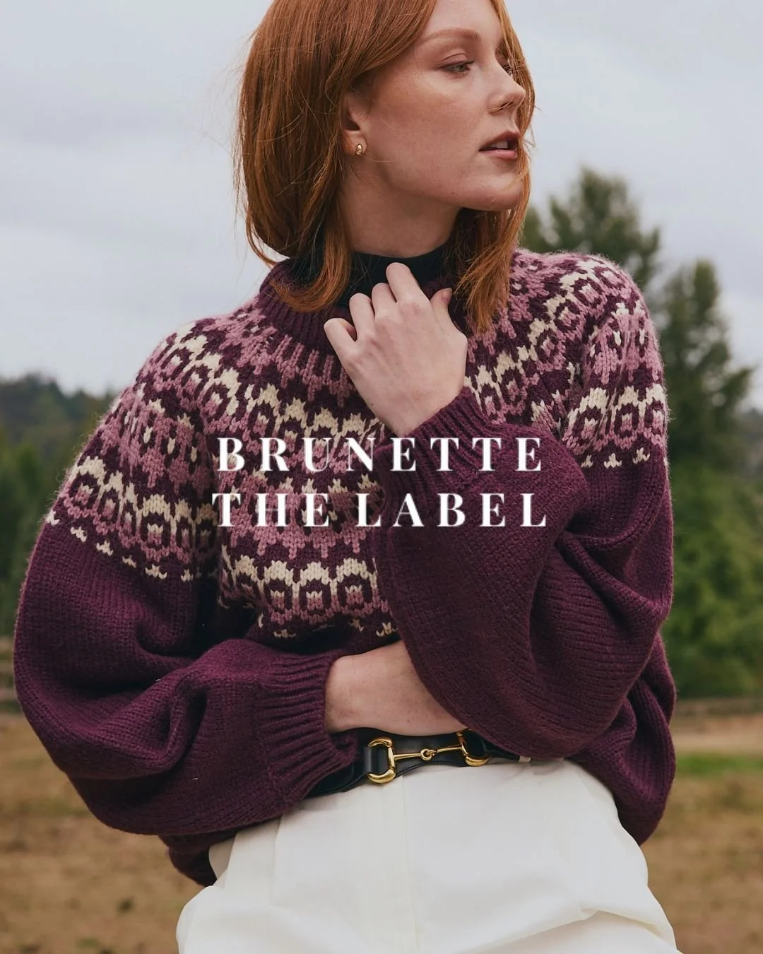 A woman with red hair wearing a dark mauve sweater with a pink and cream patterned yoke, standing outdoors with a blurred natural background.