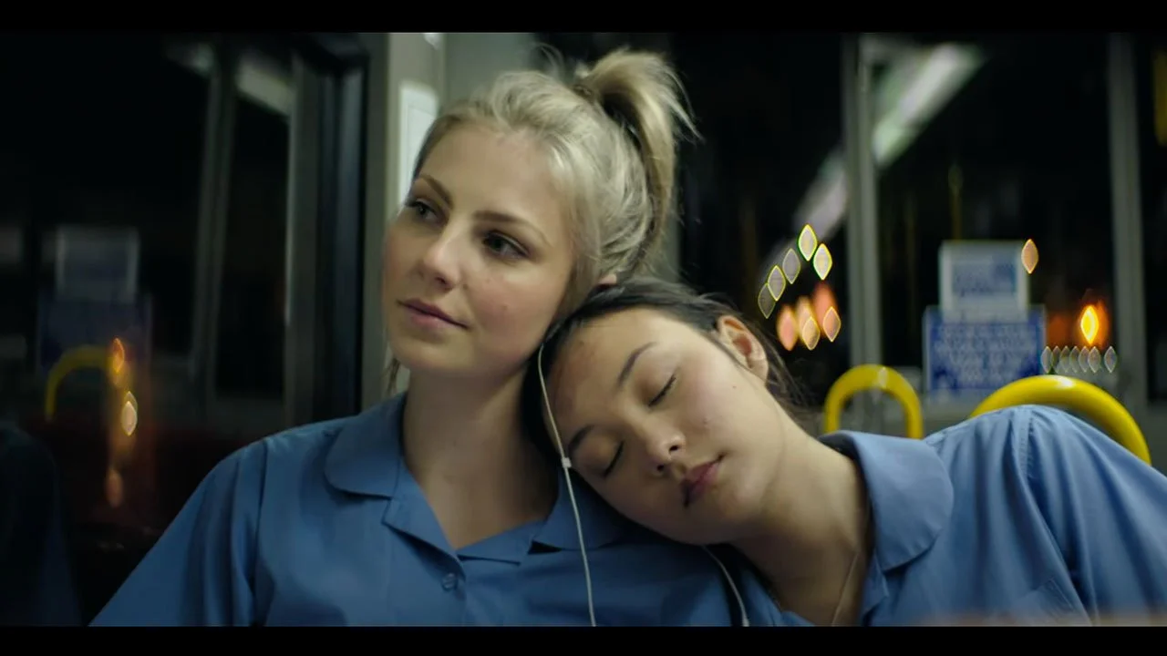 Two women wearing blue uniforms on a bus, one resting her head on the other's shoulder with her eyes closed, while the other looks slightly off to the side.